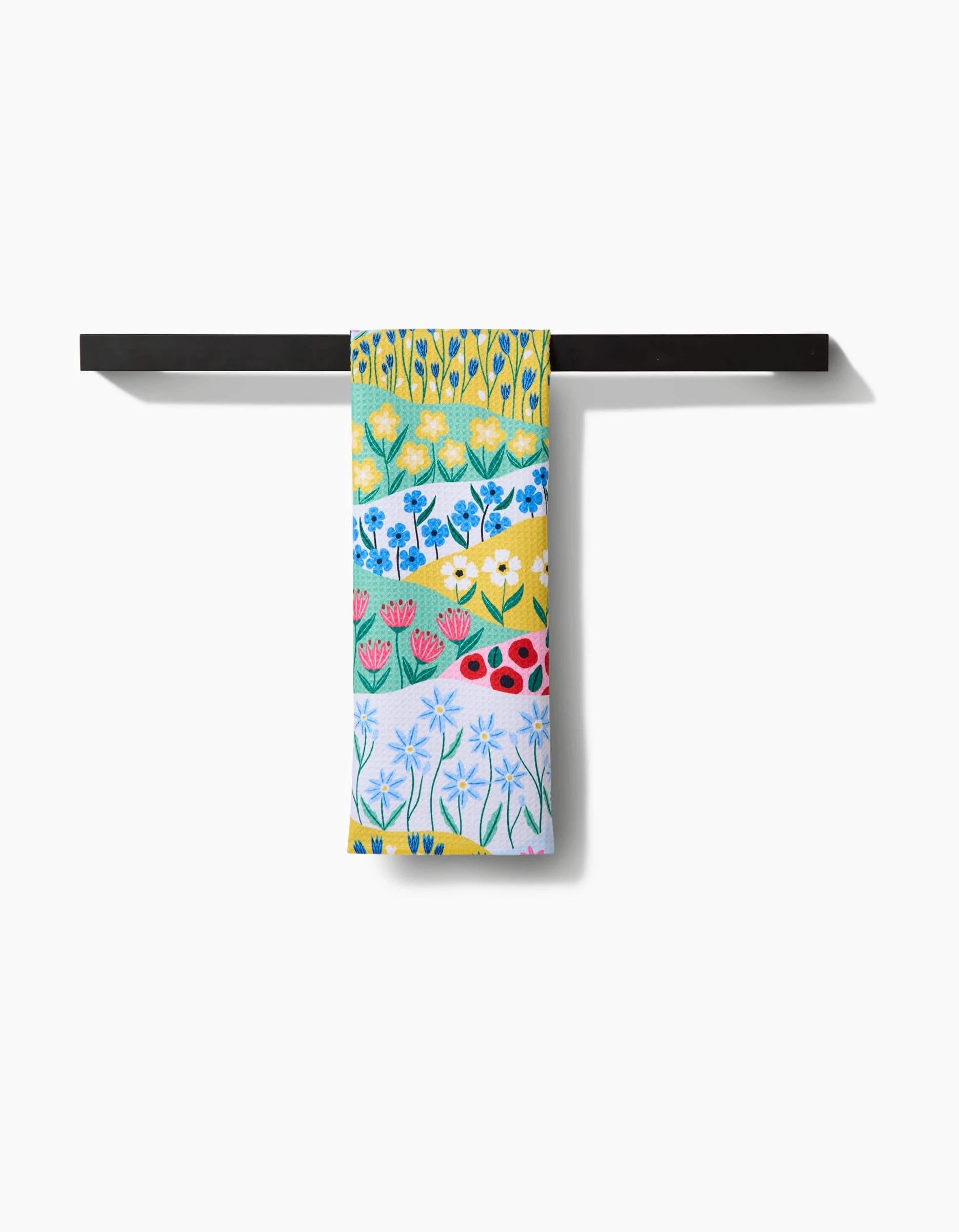 The “Spring Fields” Tea Towel by Ceyda Alasar features rows of colorful illustrated flowers and hangs neatly on a black towel rack. Crafted with Geoweave™ for superior absorbency and durability.