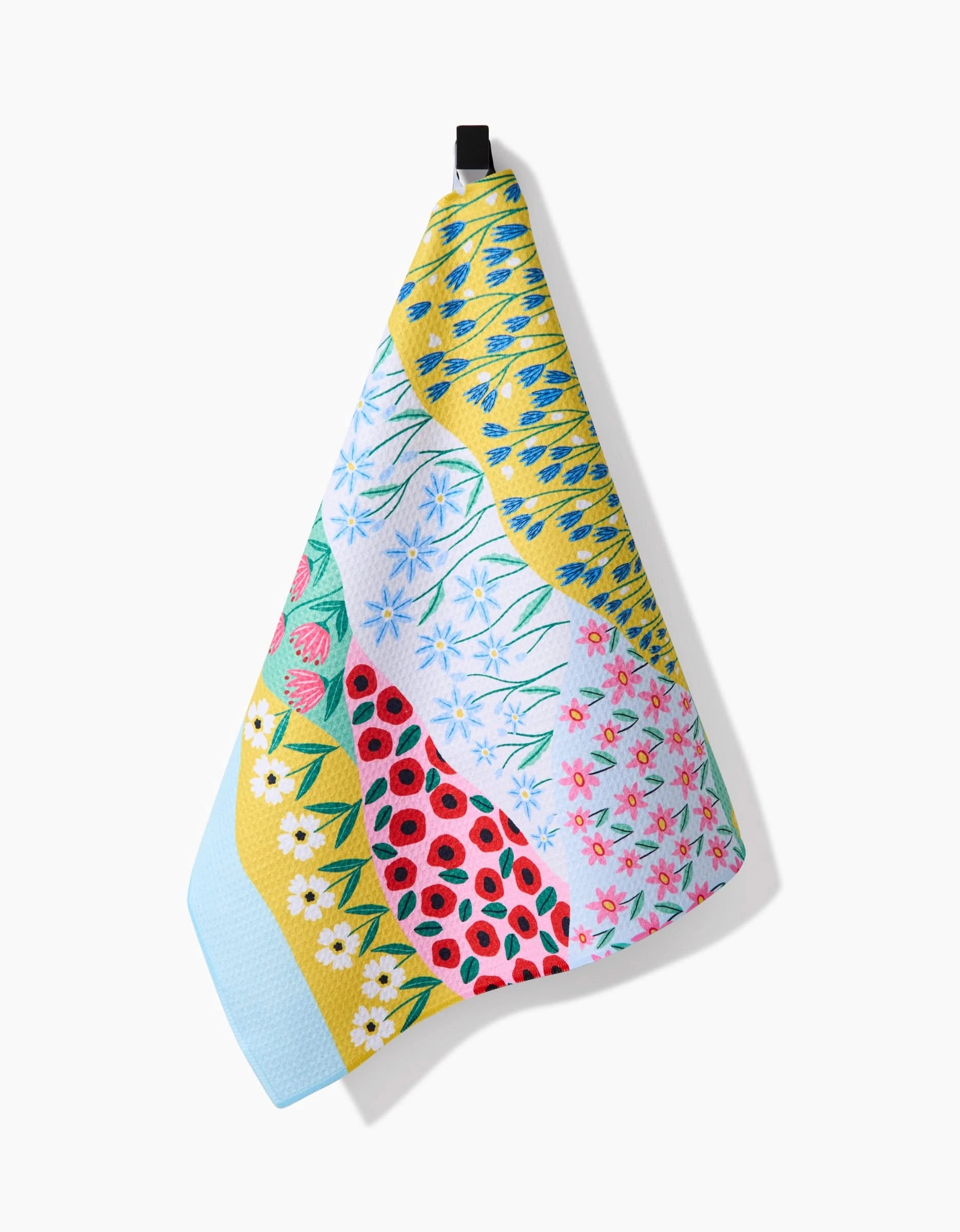 The Spring Fields tea towel by Ceyda Alasar, made from recycled materials, hangs on a hook. It displays vibrant floral patterns in yellow, blue, red, green, and white tones and casts a shadow on a white background.