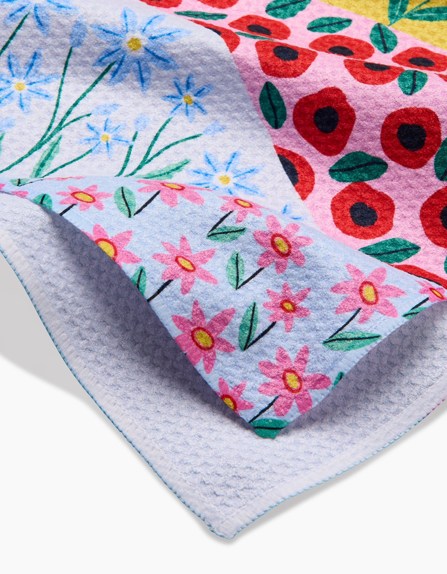 A close-up of Ceyda Alasar’s Spring Fields Geoweave™ Tea Towel, adorned with blue flowers, pink daisies, and red poppies. The partially folded towel reveals both sides and is made from recycled materials for sustainability.