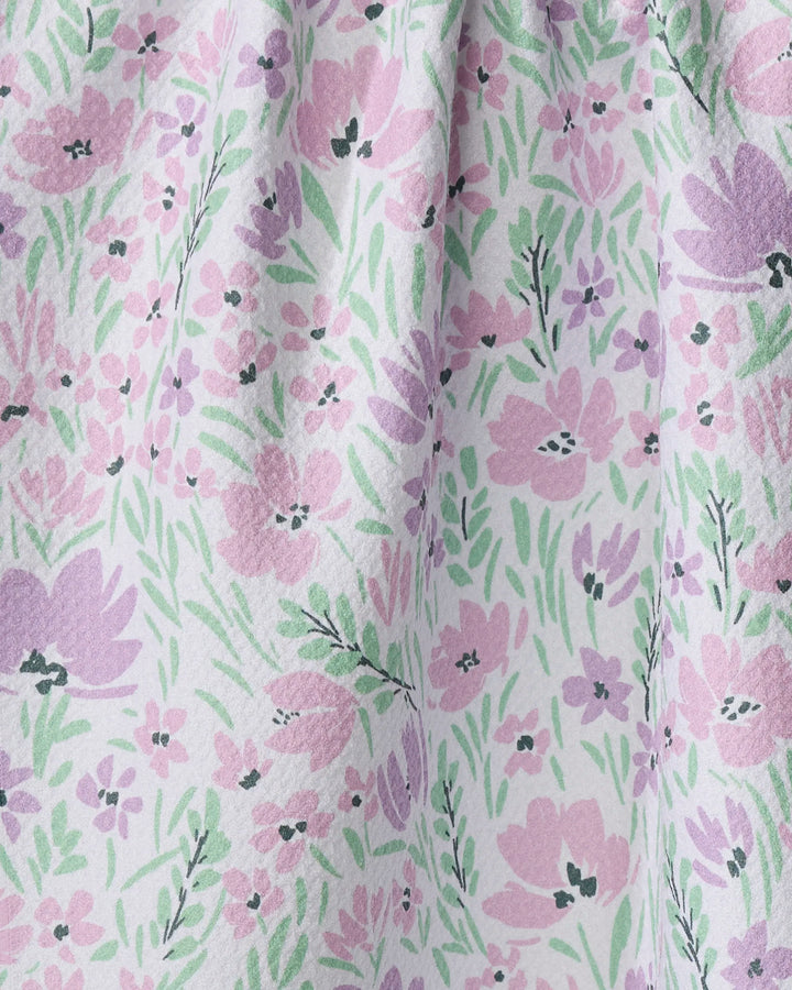 A close-up of Hufton Studio’s Spring Fleur Blooms tea towel shows a white background with pink and purple flowers, green leaves, and a slightly textured fabric made from recycled materials, gathered near the top.