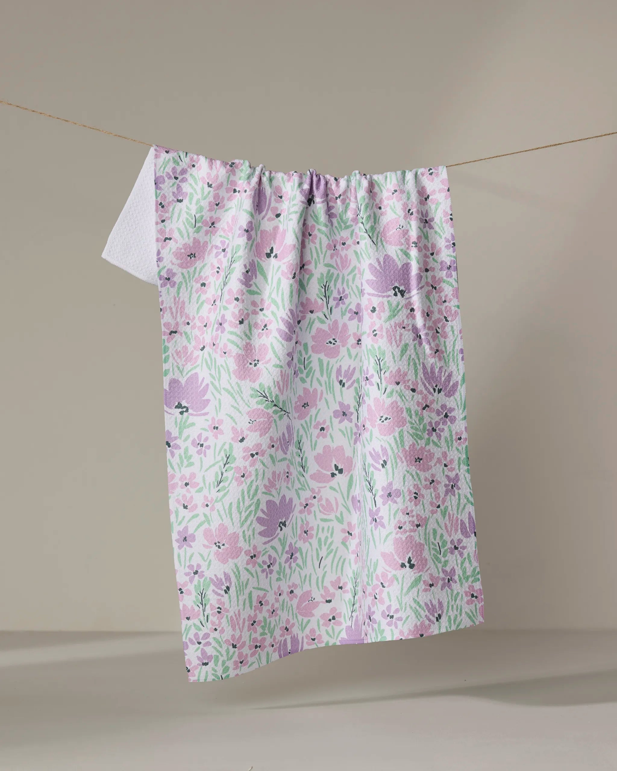 The Spring Fleur Blooms kitchen towel by Hufton Studio, featuring pink, purple, and green pastel florals and made from recycled materials, hangs on a clothesline against a neutral background.