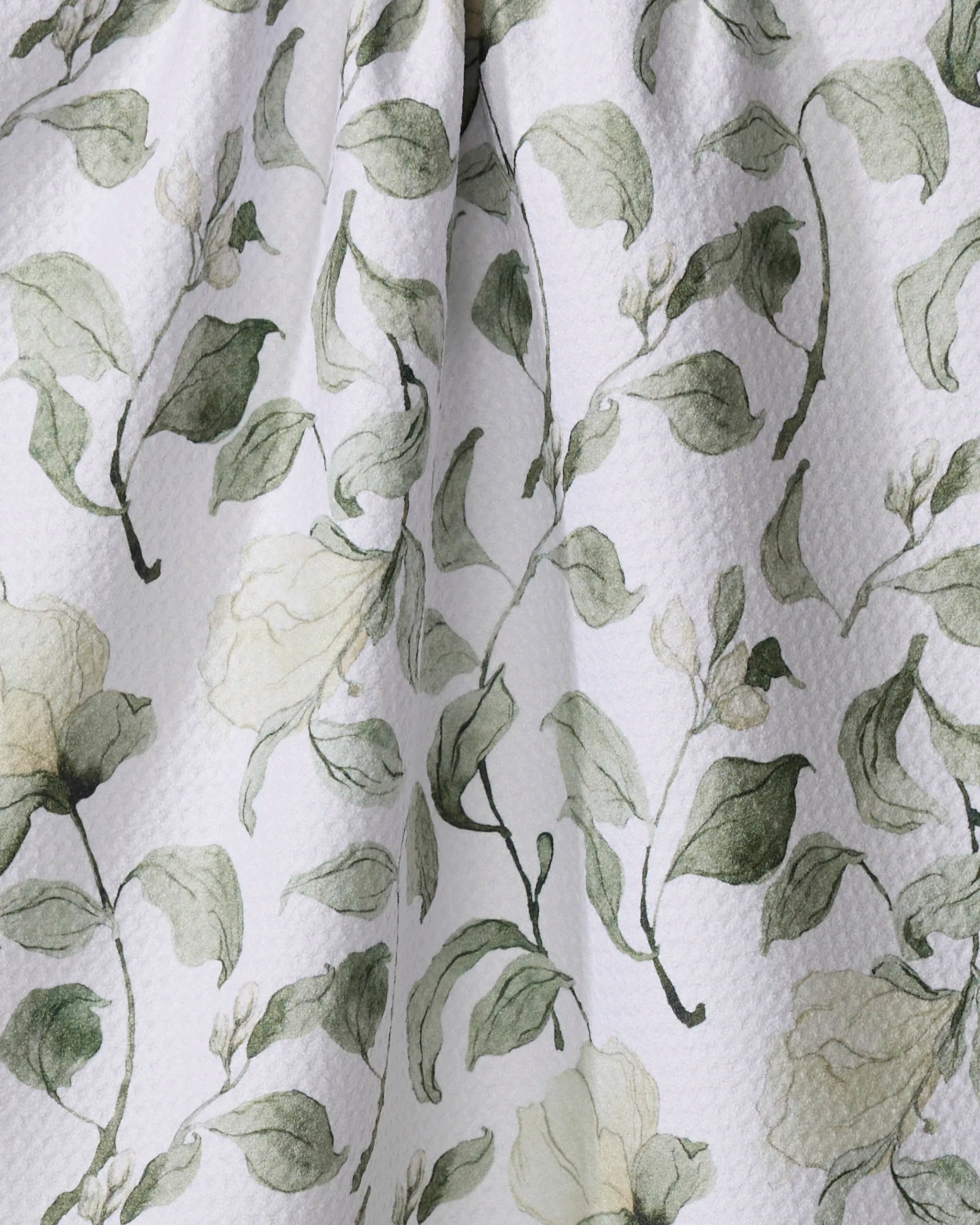 A close-up of Aubrey Fairchild’s Spring Floral Blossoms recycled kitchen towel, showcasing textured white fabric with a soft green and beige floral pattern of leaves and delicate flowers, gently draped to reveal natural folds and shadows.