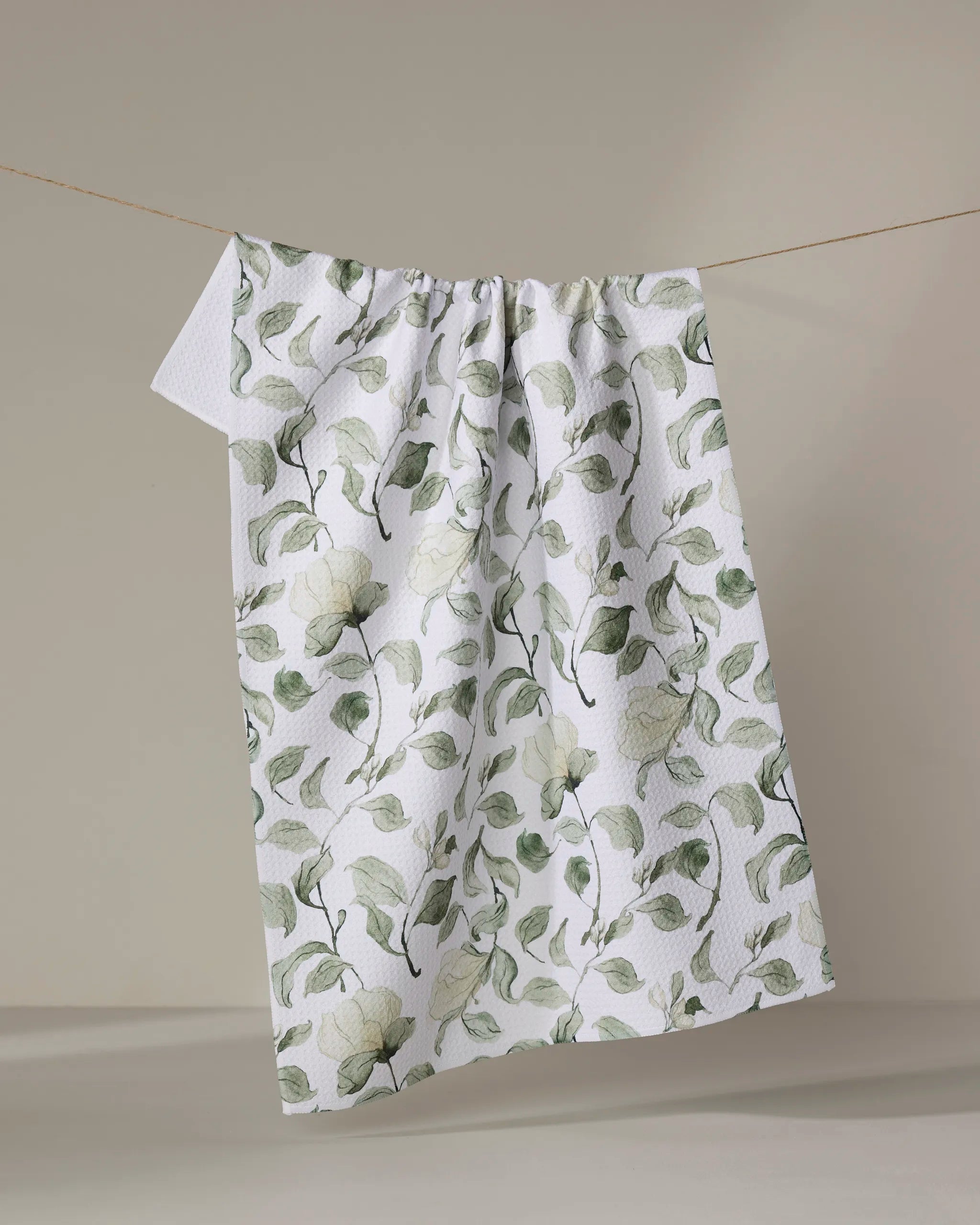The Aubrey Fairchild "Spring Floral Blossoms" recycled kitchen towel, featuring a subtle green and gray floral design, is displayed hanging on a clothesline against a neutral beige background.