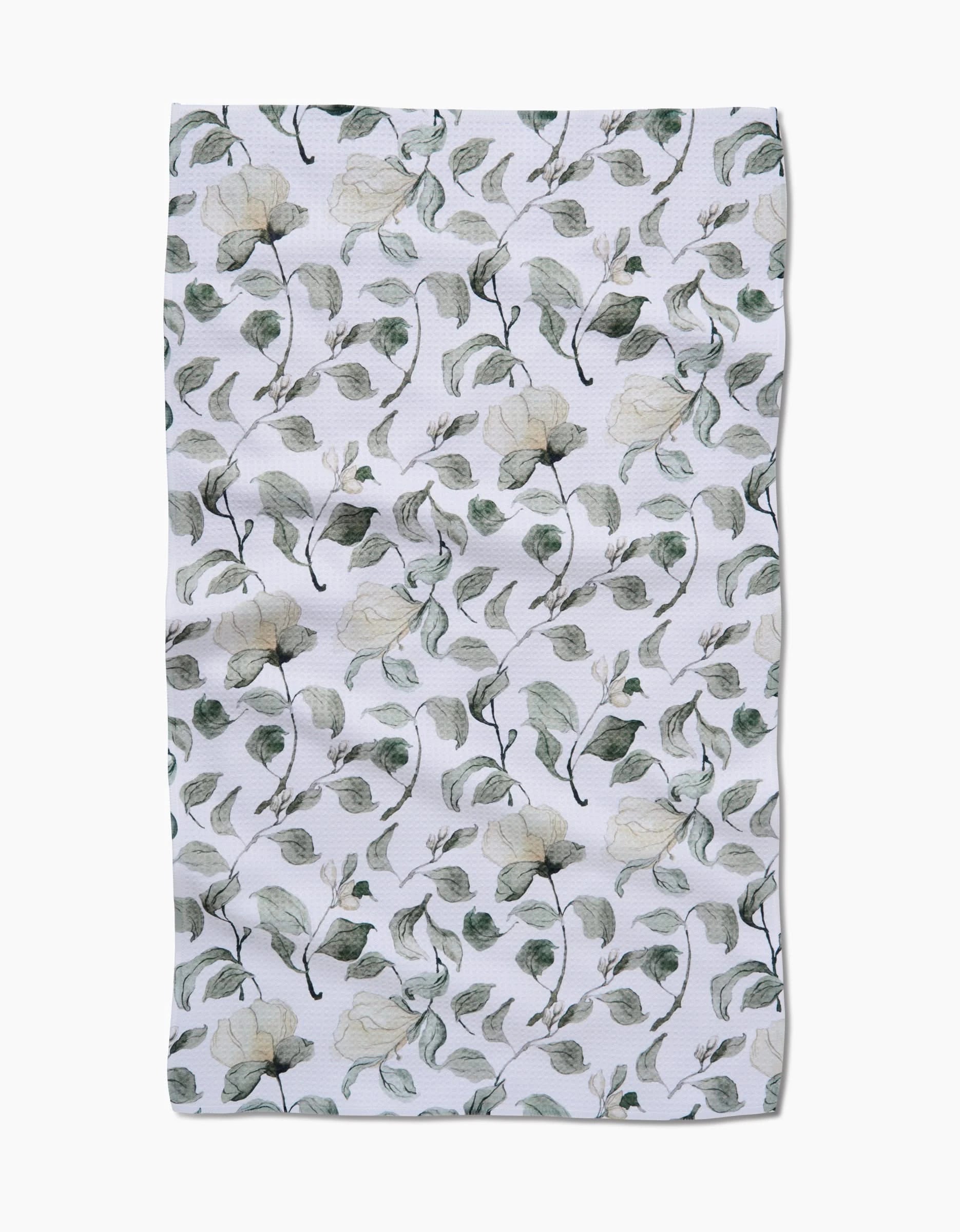 The Spring Floral Blossoms tea towel by Aubrey Fairchild features a white Geoweave fabric adorned with delicate green leaves and cream flowers. Its slightly wrinkled texture enhances the natural charm of this elegant kitchen accessory.