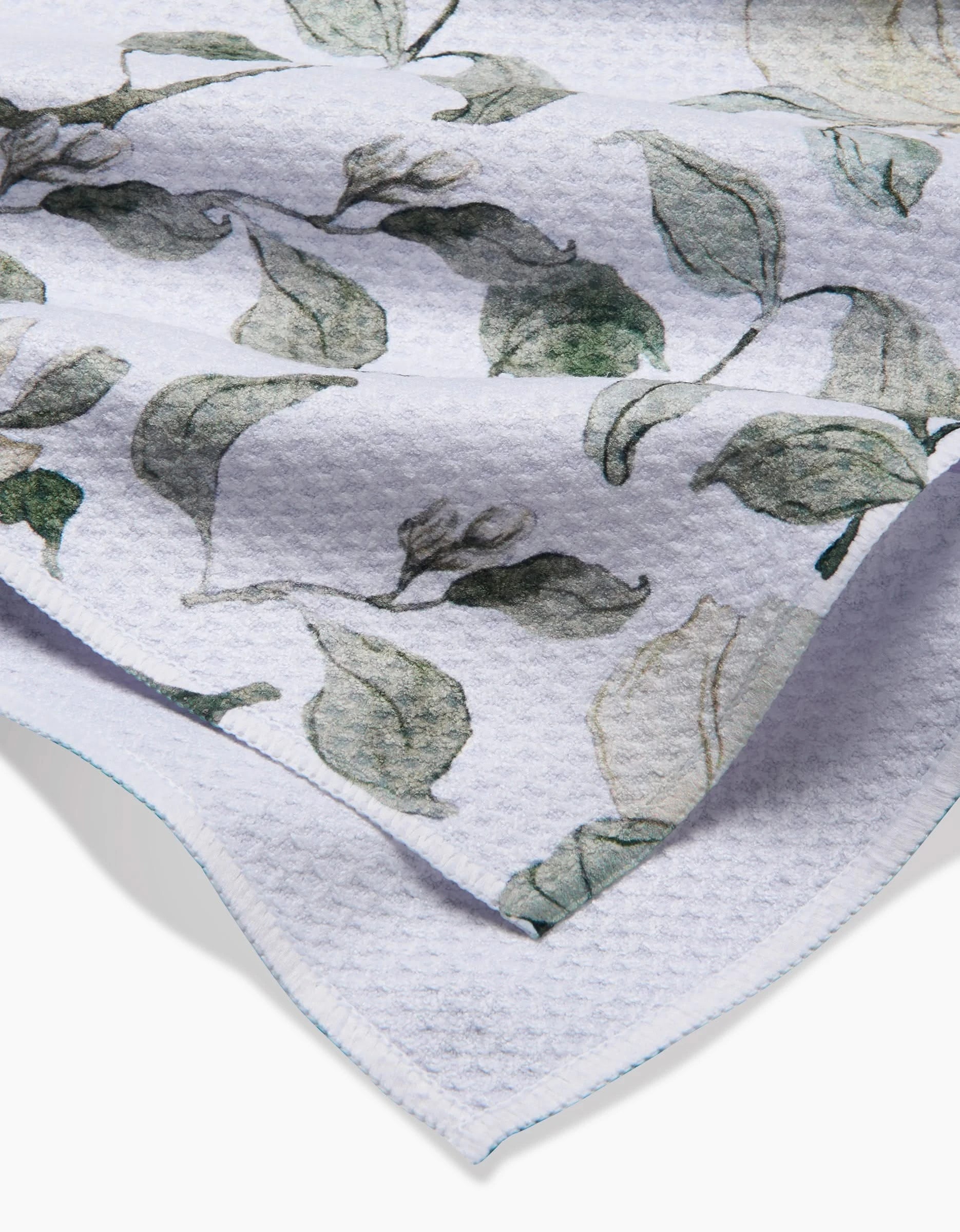 A close-up of Aubrey Fairchild’s Spring Floral Blossoms Tea Towel in white Geoweave fabric, featuring a green leafy botanical print, overlapping folds, and visible blue-stitched edges.