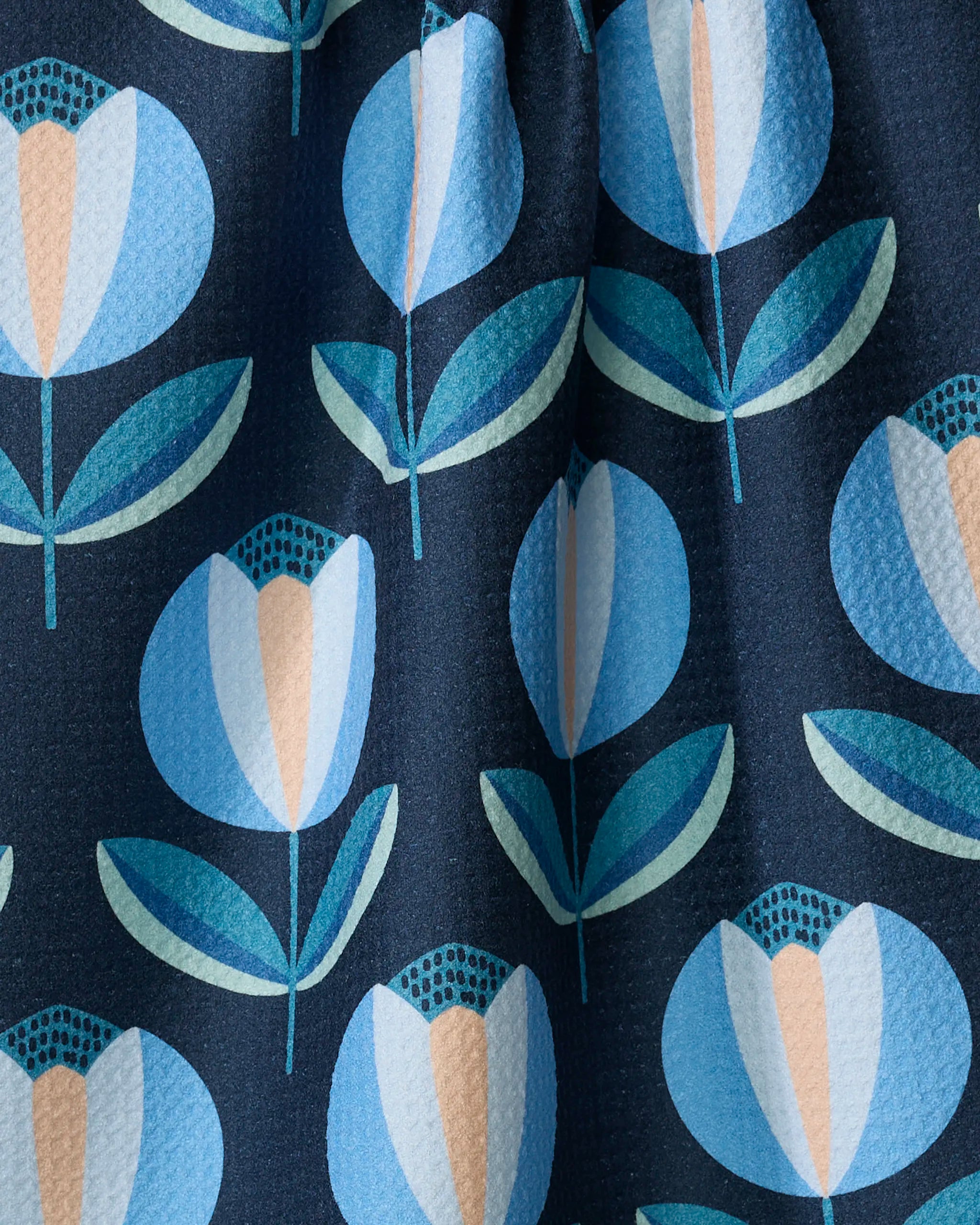 A detailed view of the Blissful Flamingo "Spring Florals" dish towel, featuring stylized blue and cream tulips with green leaves on a dark navy background. The absorbent fabric has a subtly textured finish.