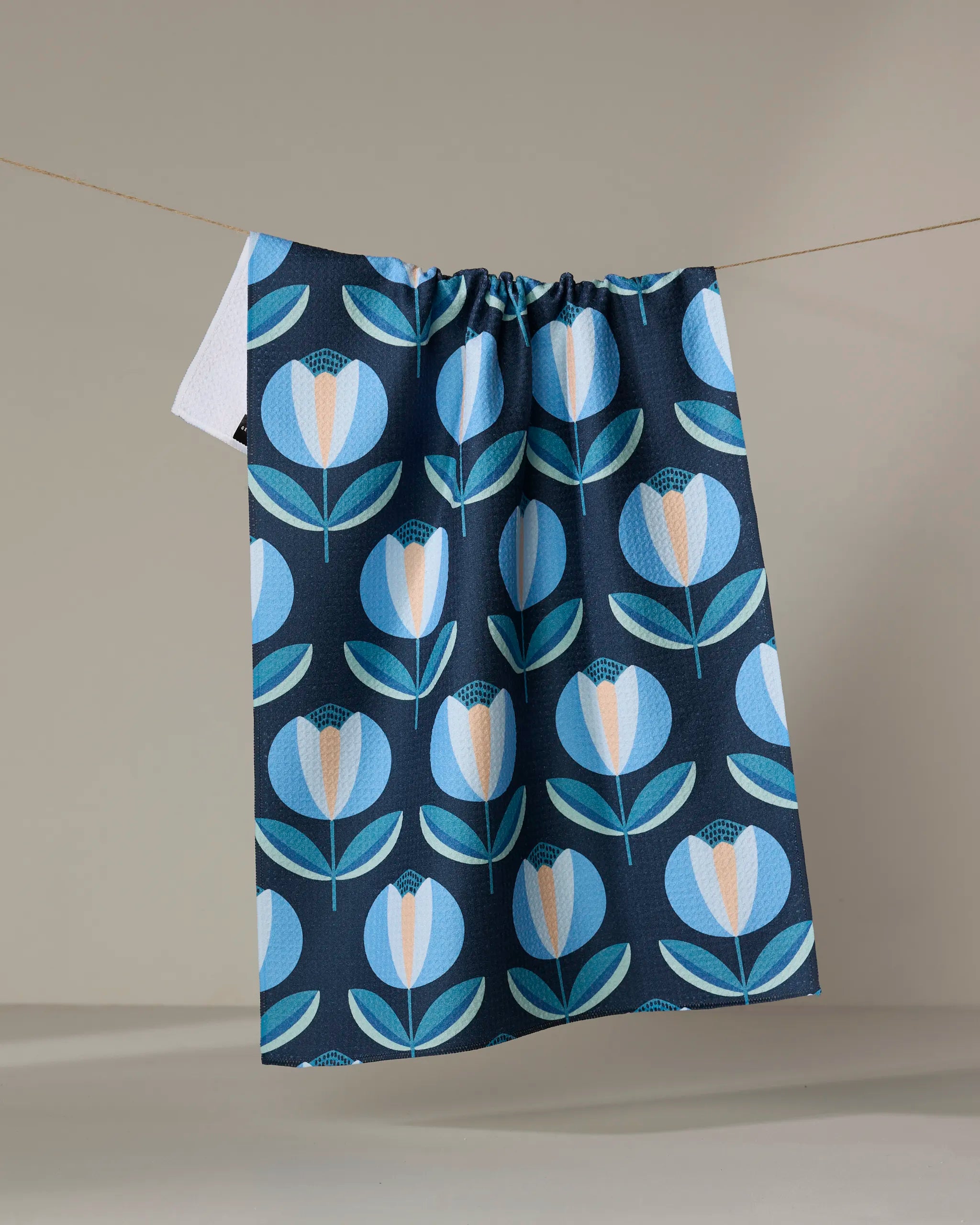 The Spring Florals dish towel by Blissful Flamingo, featuring a blue and white floral pattern, hangs on a clothesline against a neutral background.