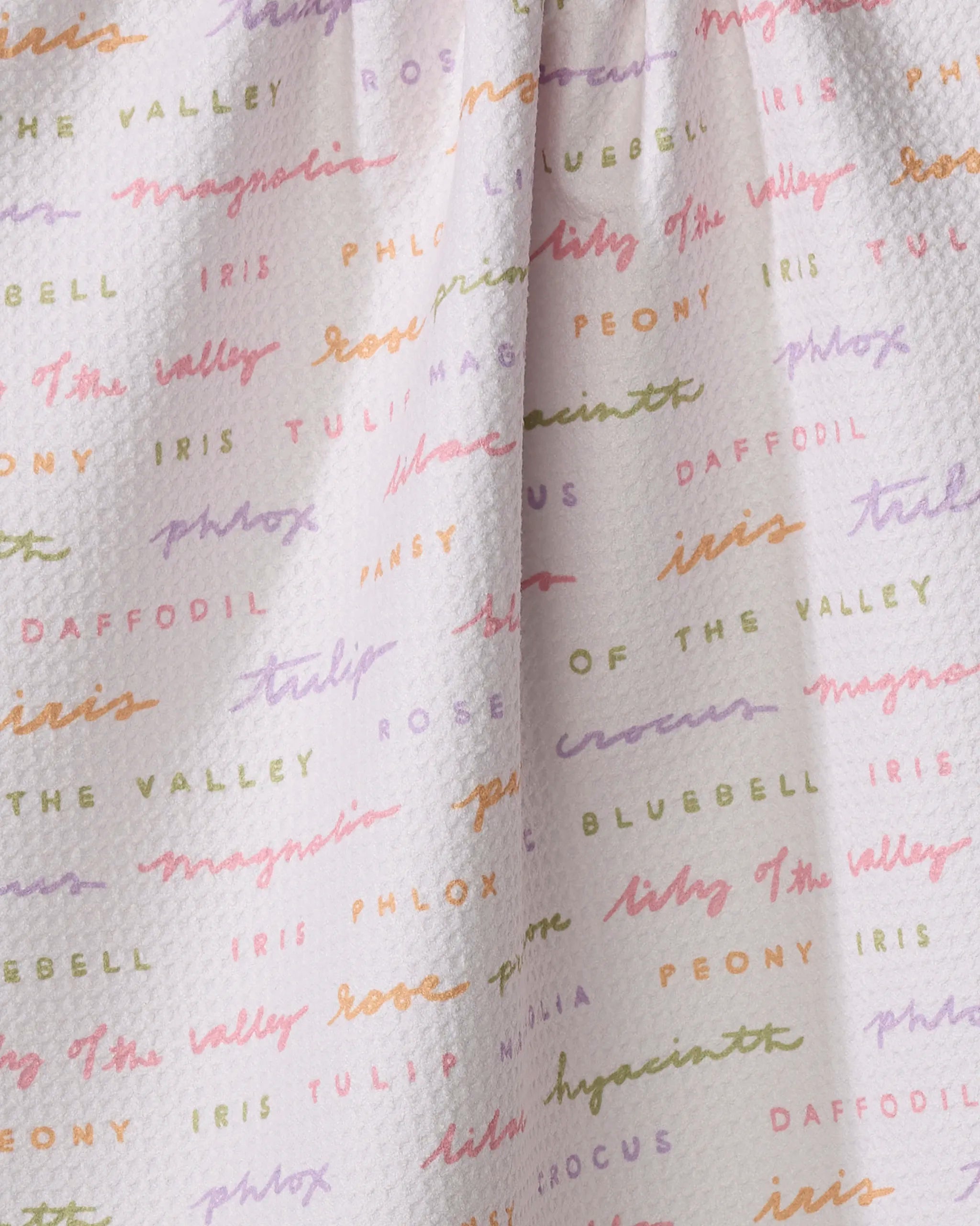 Close-up of the Kayla Ann "Spring Flowers" tea towel: a textured white dish towel featuring colorful, handwritten names of flowers in pastel shades—a charming and absorbent addition to any kitchen.