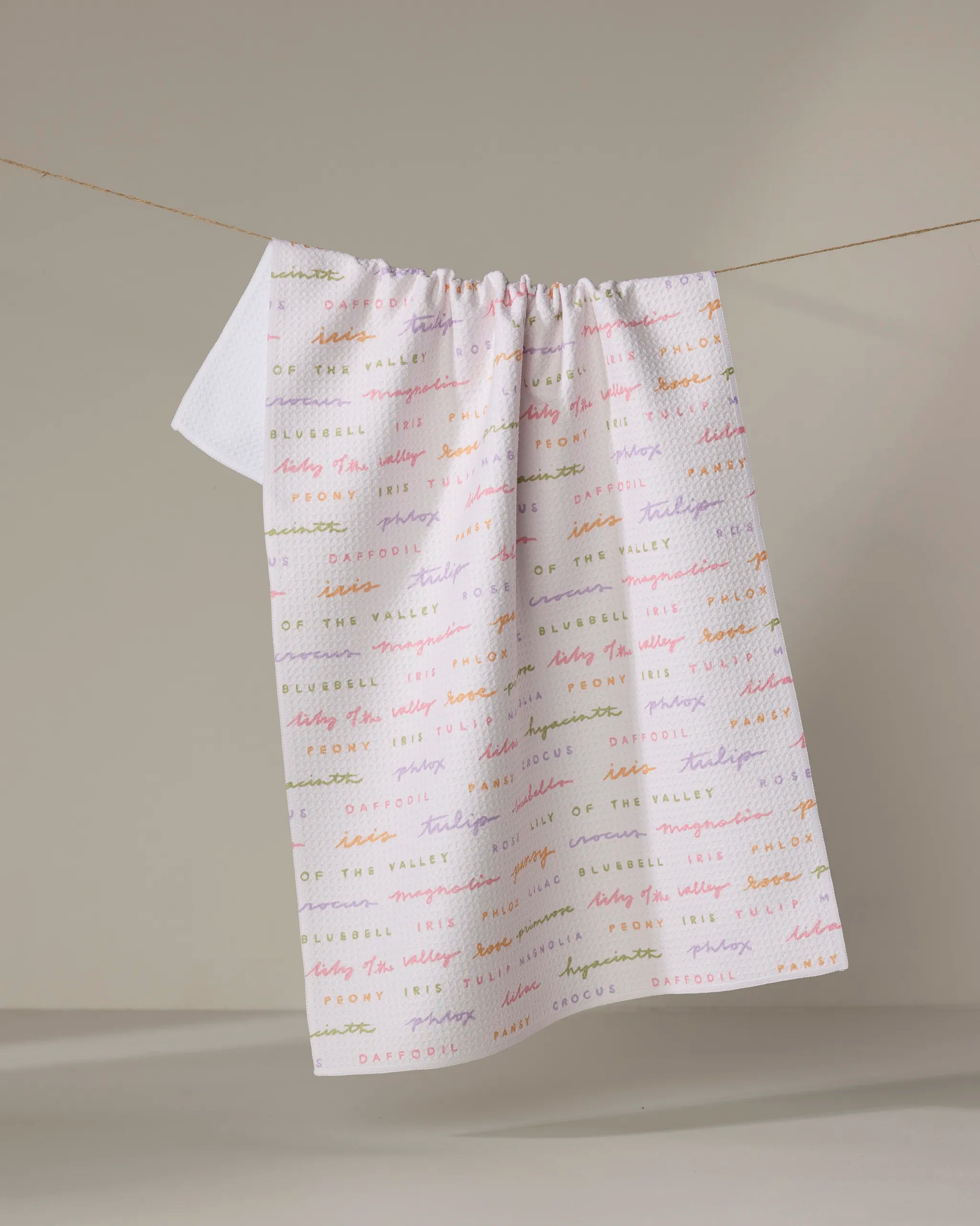 The Kayla Ann "Spring Flowers" tea towel, with pastel flower names, hangs over a string against a neutral background—a charming and absorbent touch to brighten any kitchen.