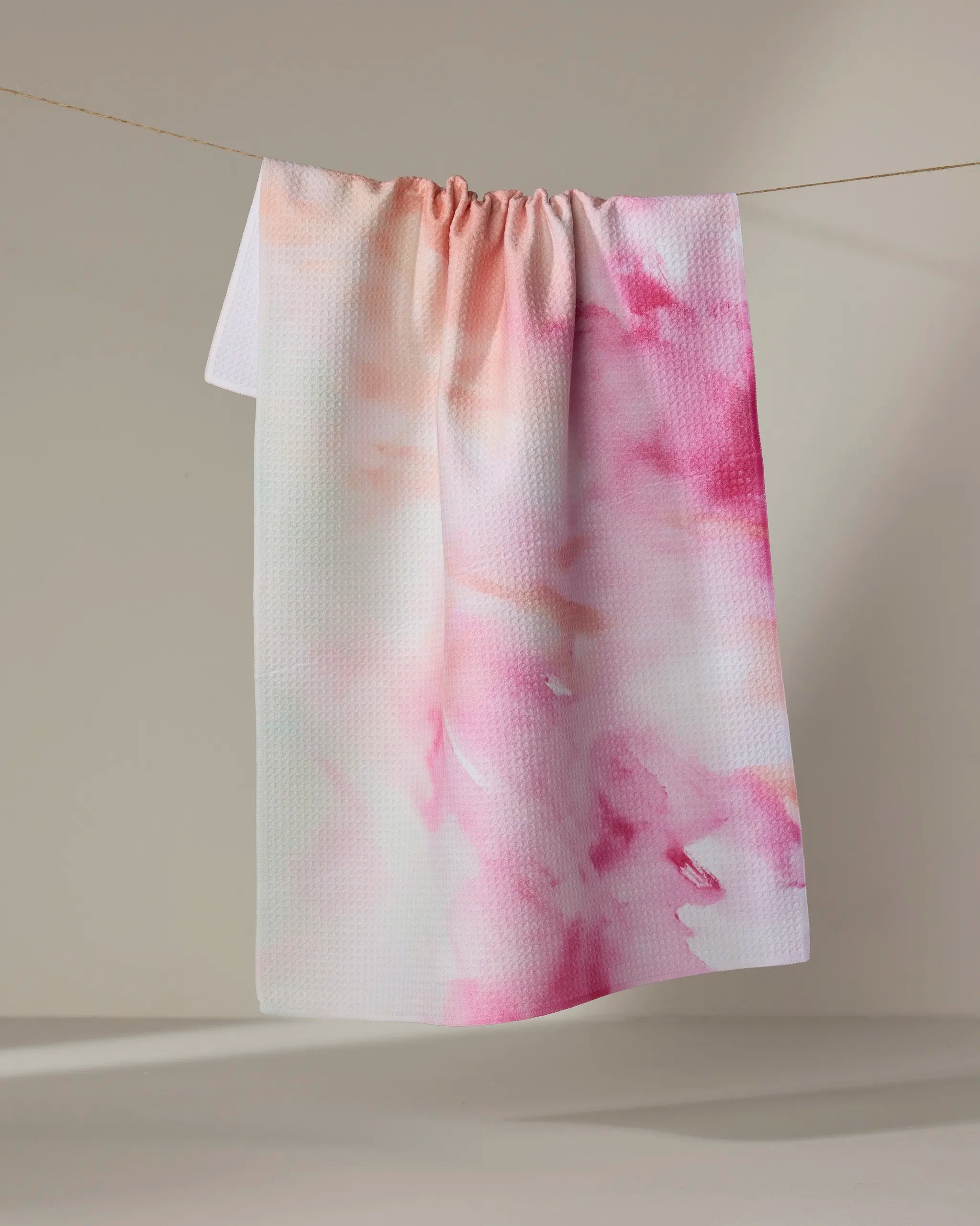 The Stefanie Bales "Spring Garden" towel, featuring a pink and white watercolor pattern, hangs on a clothesline against a neutral background.