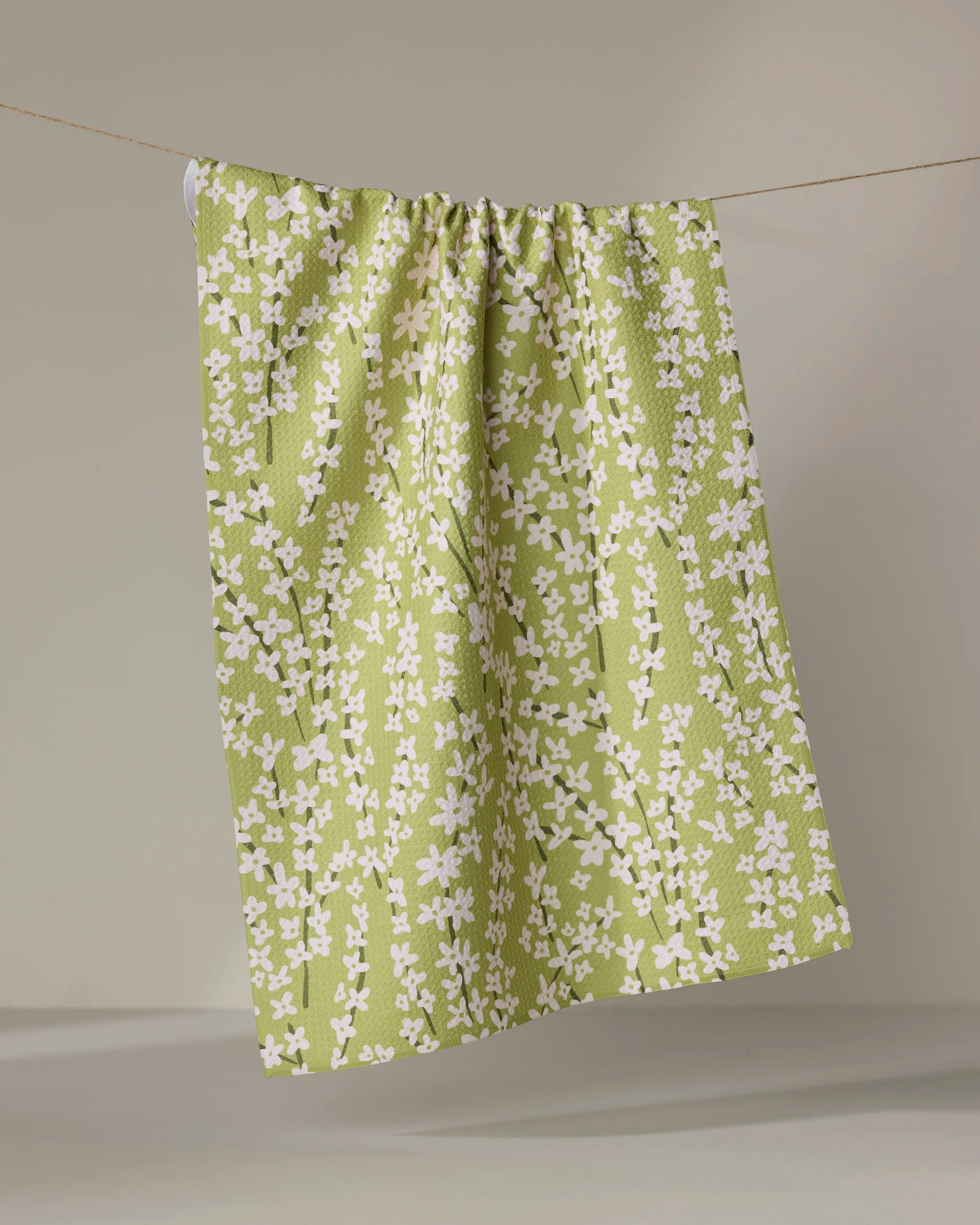 The Kayla Ann Spring Green Forsythia tea towel, featuring a white floral pattern, hangs neatly from a clothesline against a neutral background.