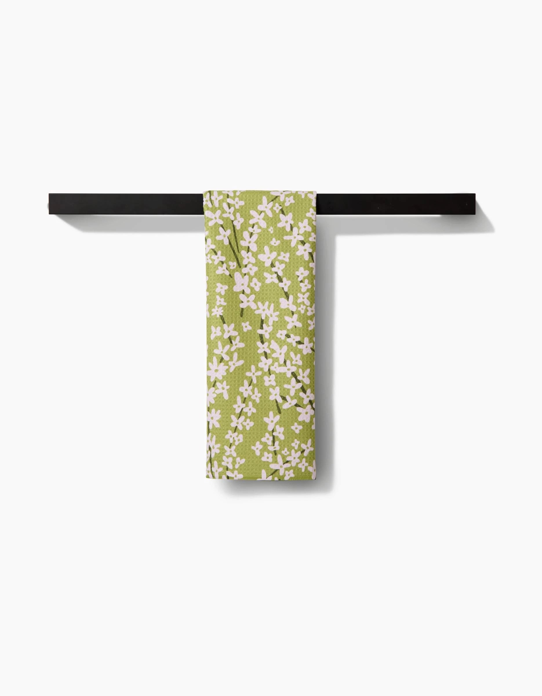 The Kayla Ann Spring Green Forsythia towel, made from ultra-absorbent Geoweave fabric and featuring a white floral pattern, hangs neatly on a black bar against a white background.