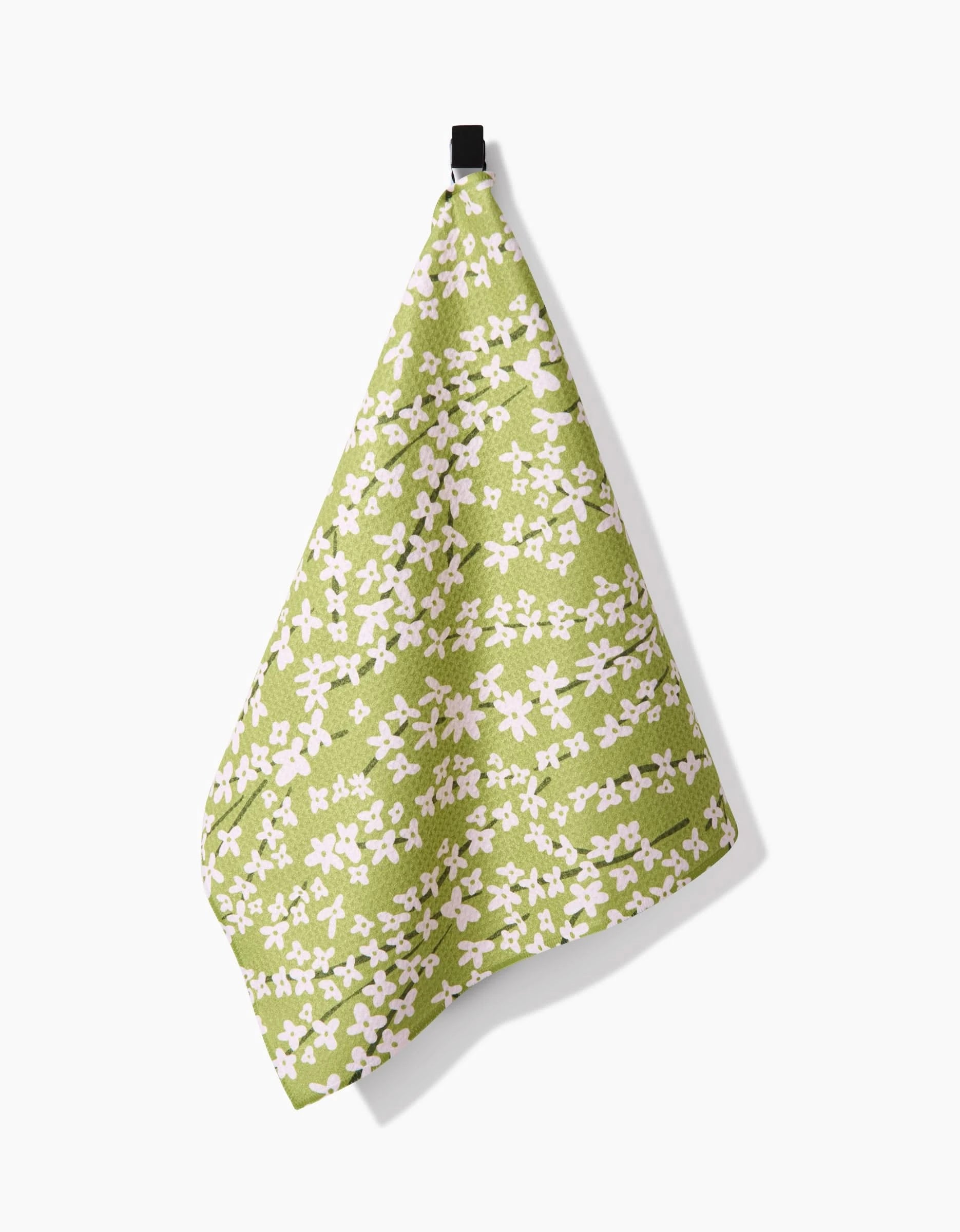 The Kayla Ann Spring Green Forsythia dish towel, made from recycled materials and featuring small white flowers, hangs on a black hook against a white background.