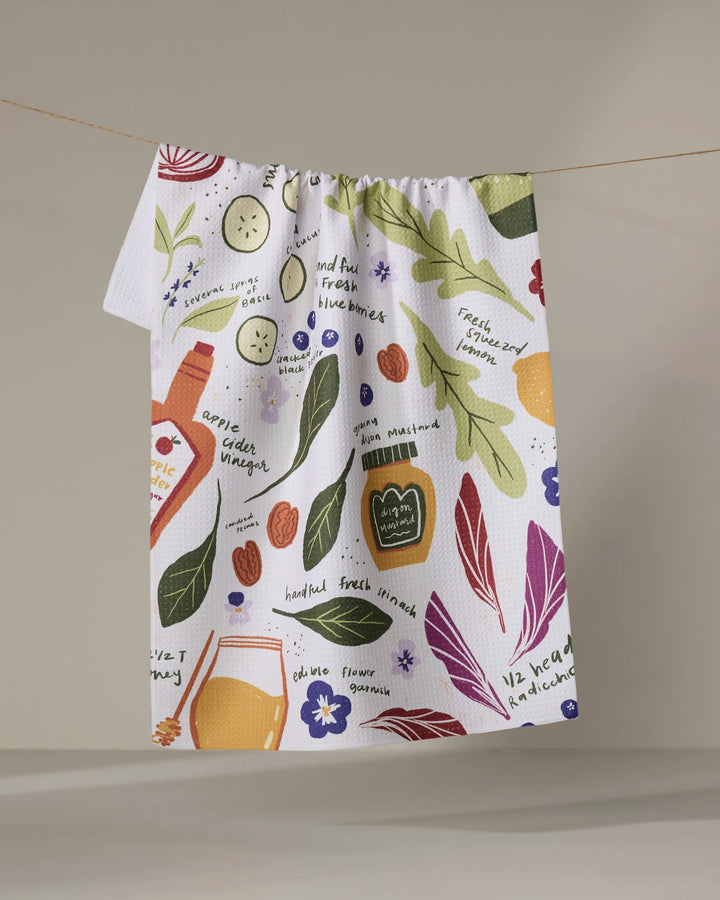 The Spring Harvest Salad tea towel by Janna Sue Design hangs on a line, featuring vibrant illustrations of veggies, fruits, herbs, and recipe notes in multiple colors on a white background.