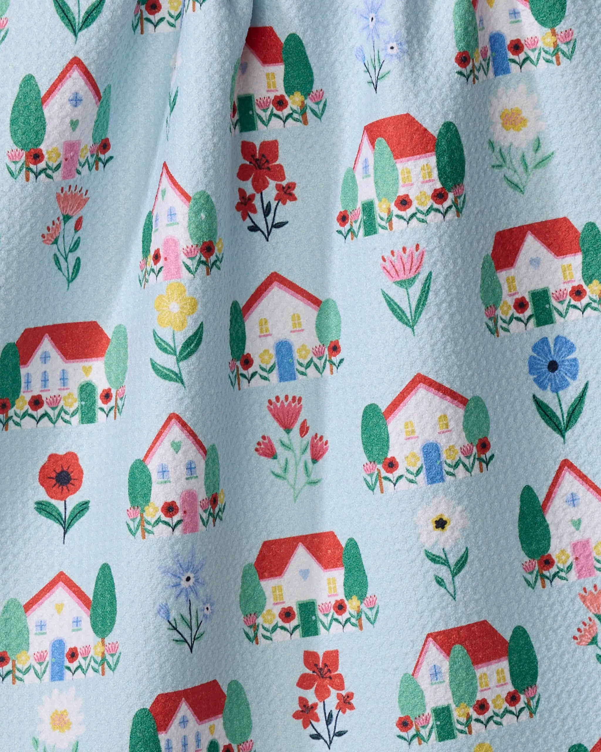 The "Spring Houses" tea towel by Ceyda Alasar is quick-drying and features small, colorful houses with red roofs, trees, and bright flowers on a light blue textured background.