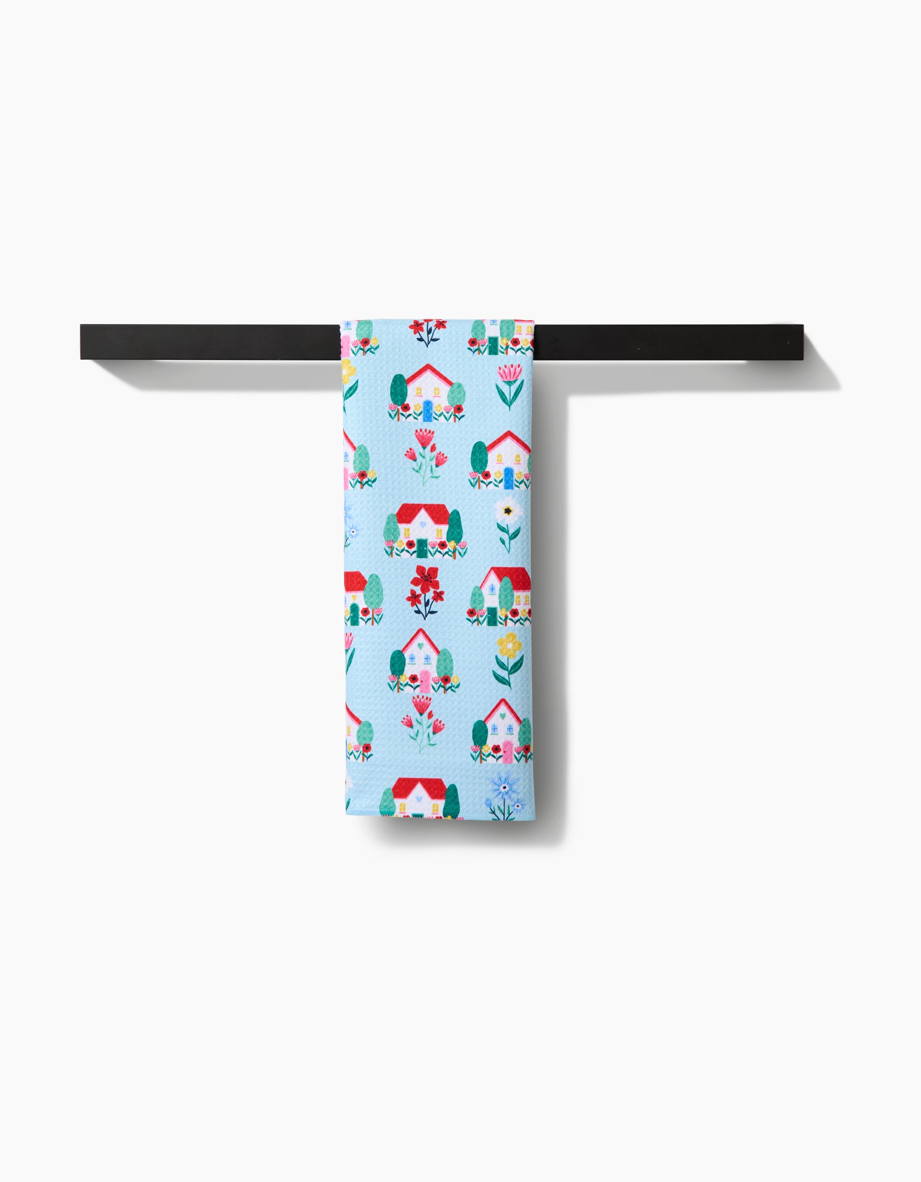 Ceyda Alasars Spring Houses tea towel, made from recycled materials, displays colorful house and flower illustrations. Shown in light blue, it hangs neatly over a black rack with a white background.