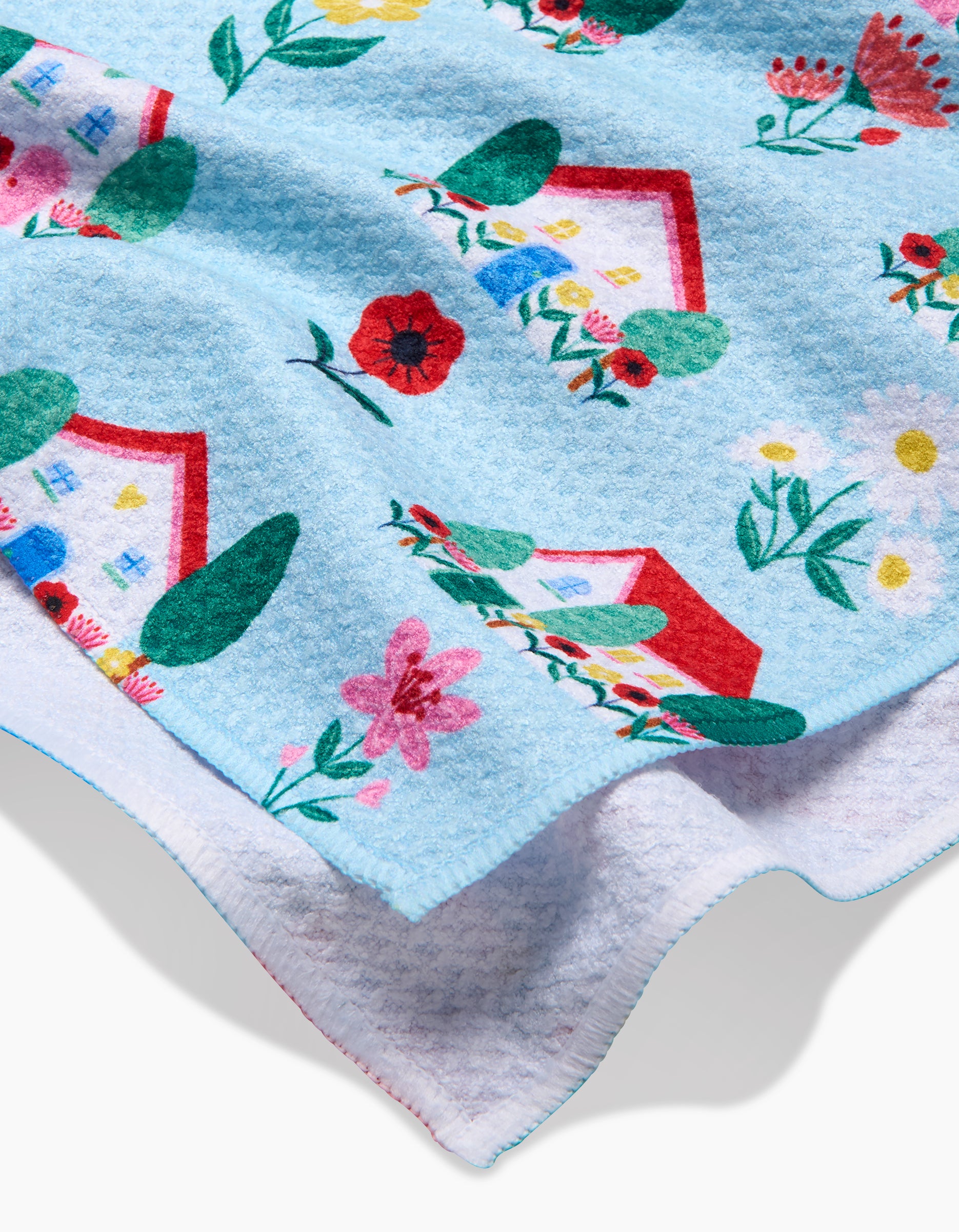 The Ceyda Alasar Spring Houses blanket features soft blue Geoweave fabric, colorful prints of houses, flowers, and greenery, a white reverse side, multicolored embroidered edges, and is crafted from recycled materials for sustainability.