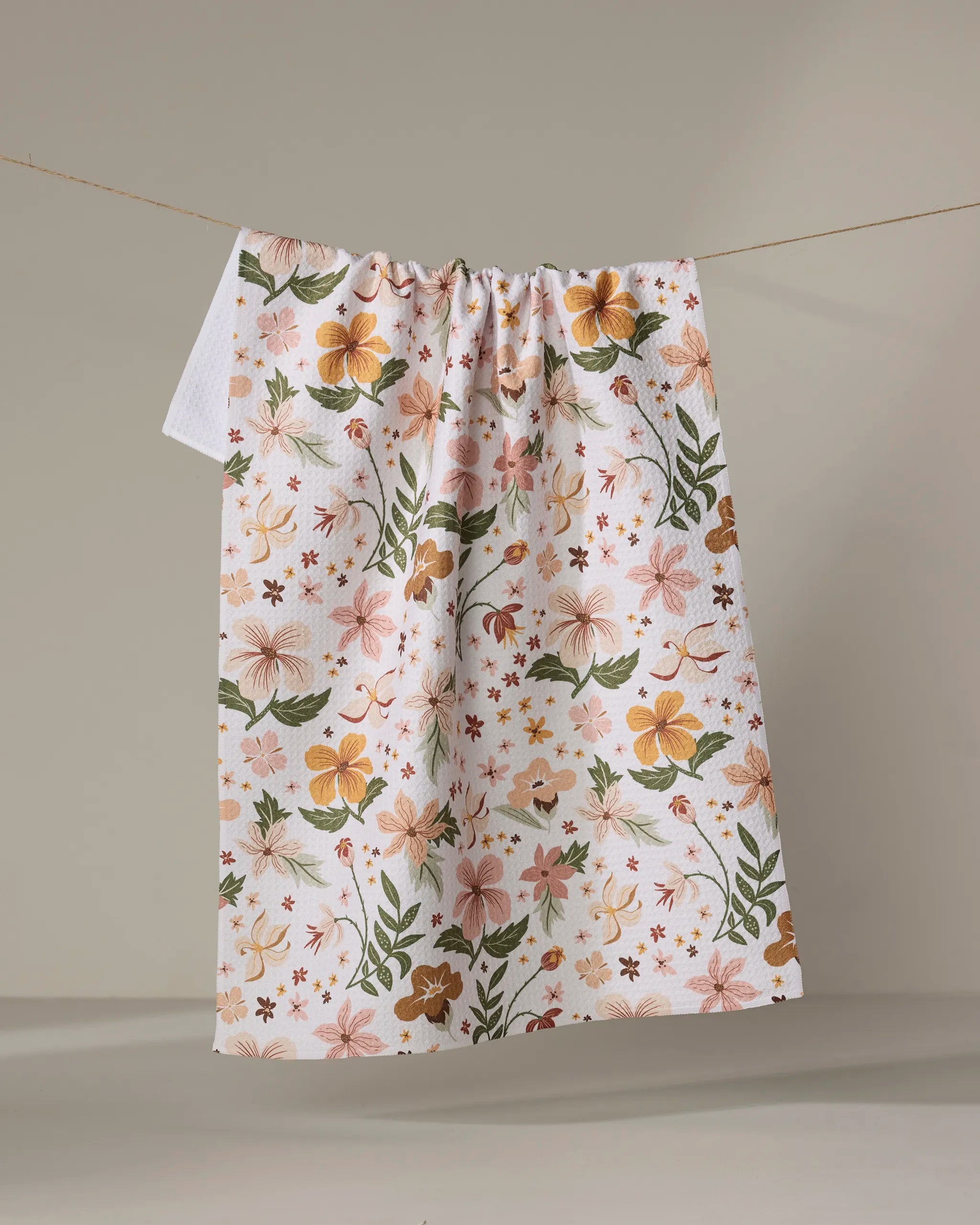 The "Spring In Devon" tea towel by Danelys Sidron features a vibrant floral pattern in orange, pink, and green on white, hanging on a clothesline against a neutral background.