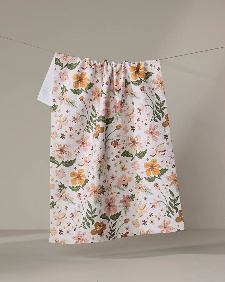 The "Spring In Devon" tea towel by Danelys Sidron features a vibrant floral pattern in orange, pink, and green on white, hanging on a clothesline against a neutral background.