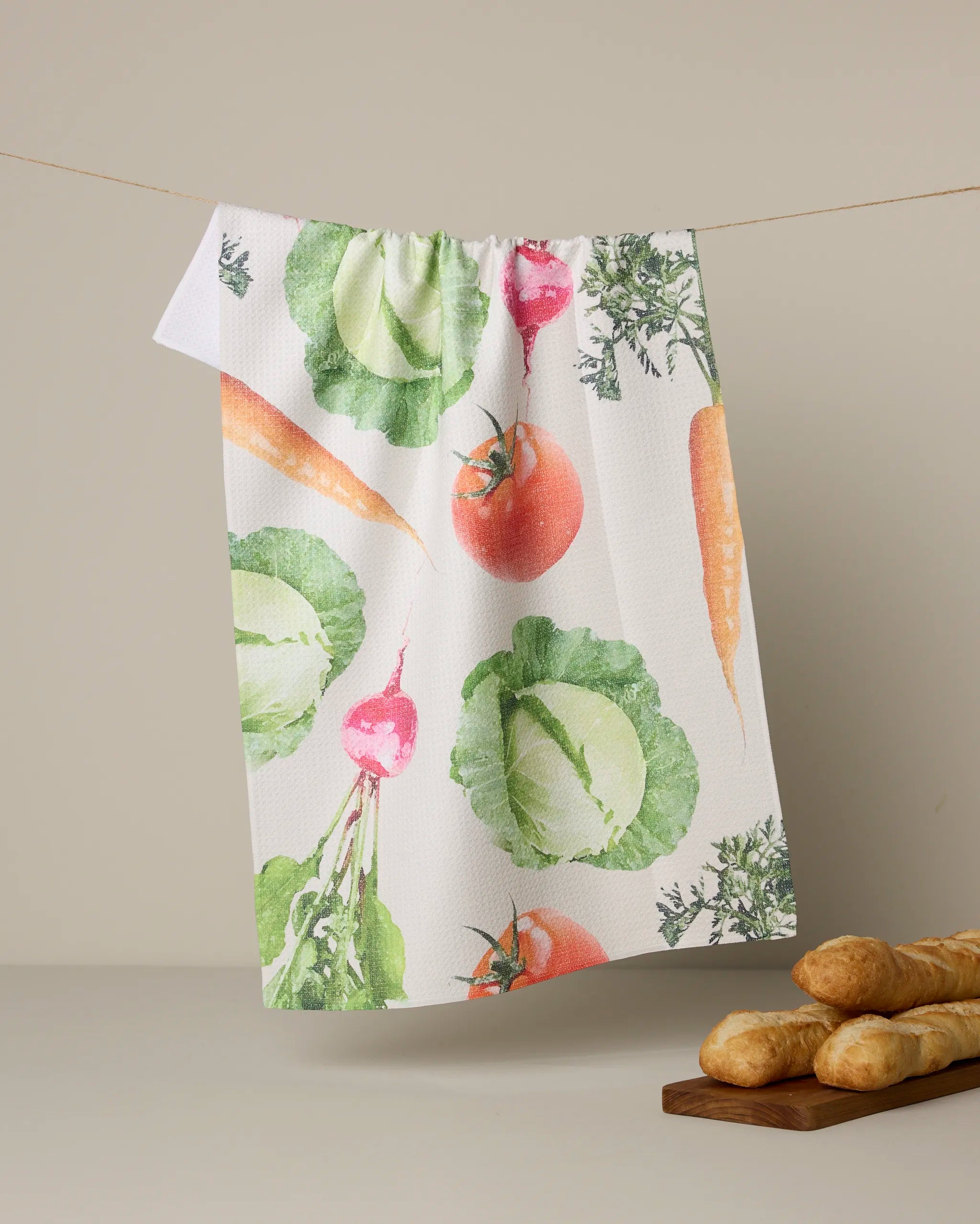 The Geometry “Spring Produce” tea towel, featuring colorful illustrations of carrots, tomatoes, lettuce, and radishes on a plain beige background, hangs above baguettes and adds quick-drying charm to any kitchen.