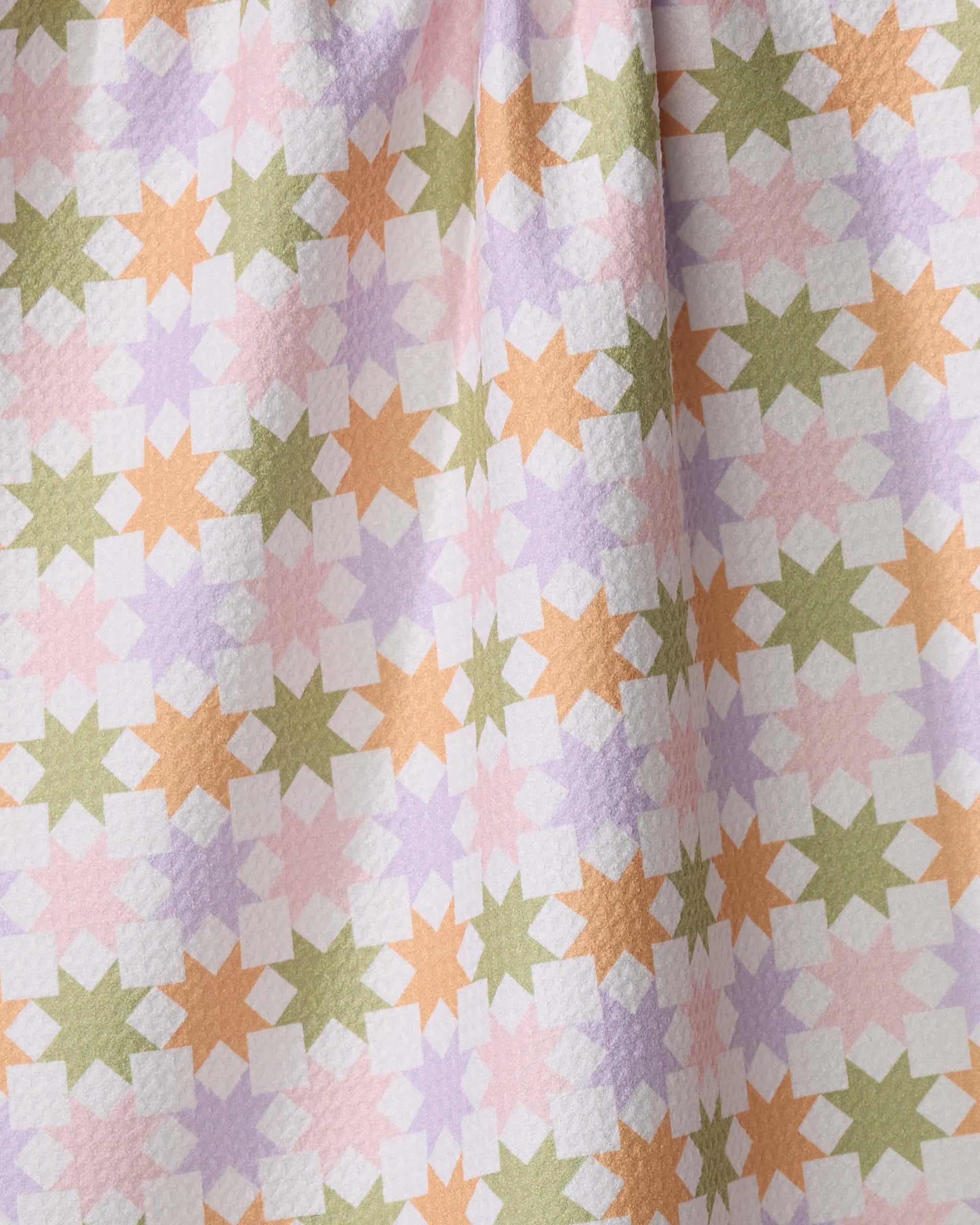 A close-up of Kayla Ann's Spring Quilt, featuring geometric star shapes in orange, green, lavender, and white on recycled fabric. The quilt has visible folds and a slightly textured surface arranged in a repeating pattern.
