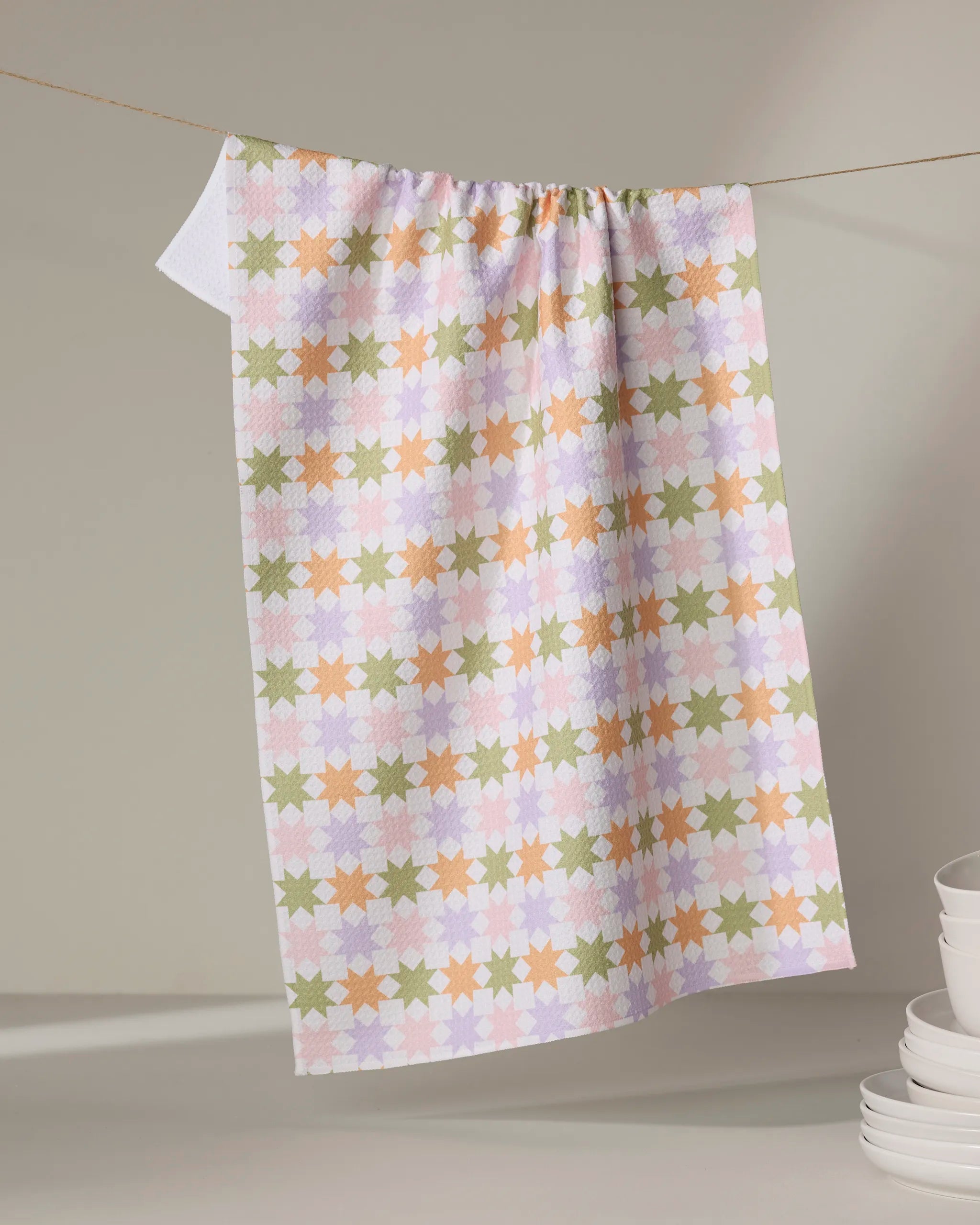 The Kayla Ann "Spring Quilt" tea towel features a pastel star pattern in orange, green, and lavender on recycled fabric. Hanging on a string, it appears with stacked white dishes in the lower right against a light background.