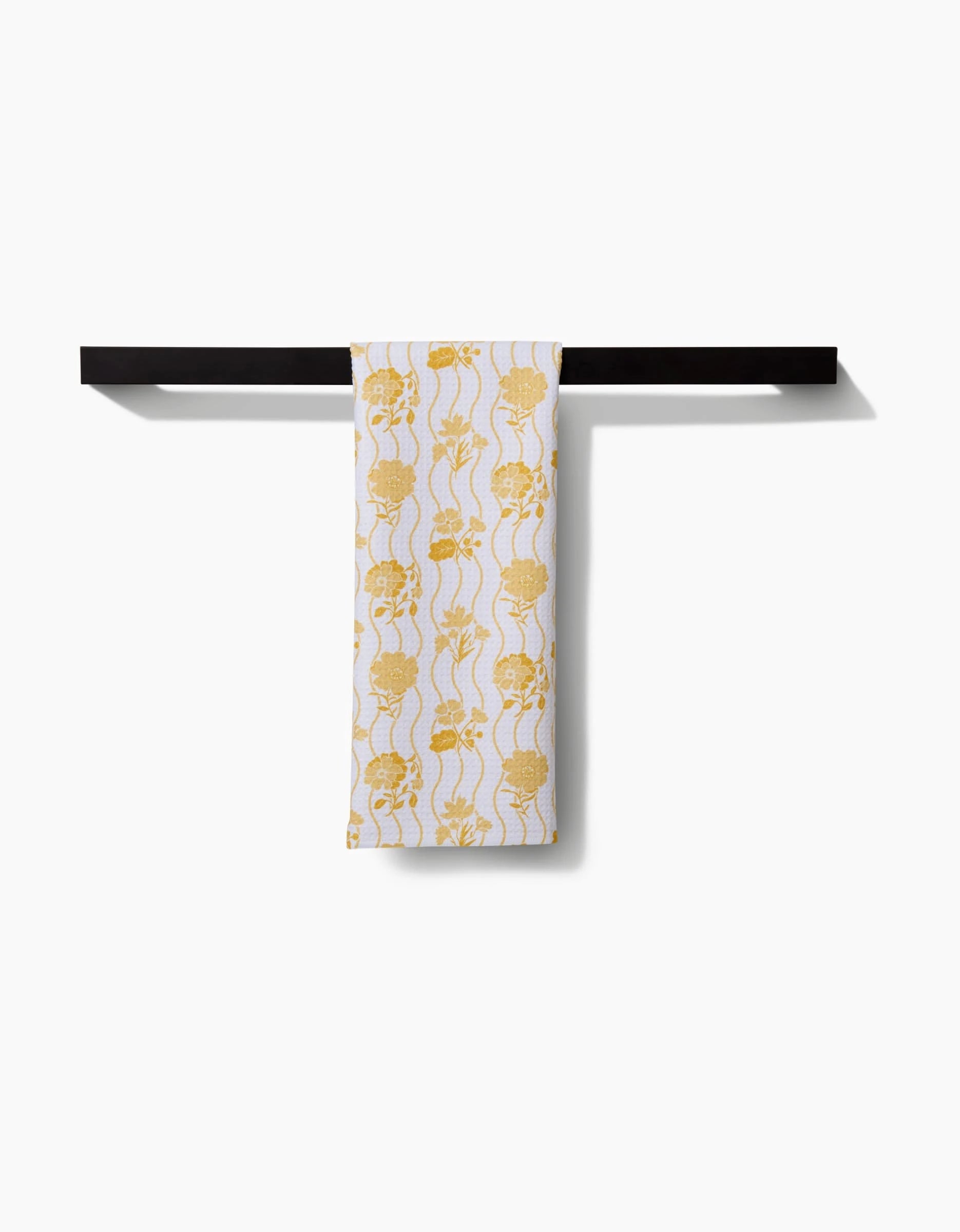 A Danelys Sidron Spring Wave tea towel with a yellow floral pattern hangs on a black rack against a white wall. Made from Geoweave fabric, it blends absorbency and style for your kitchen décor.