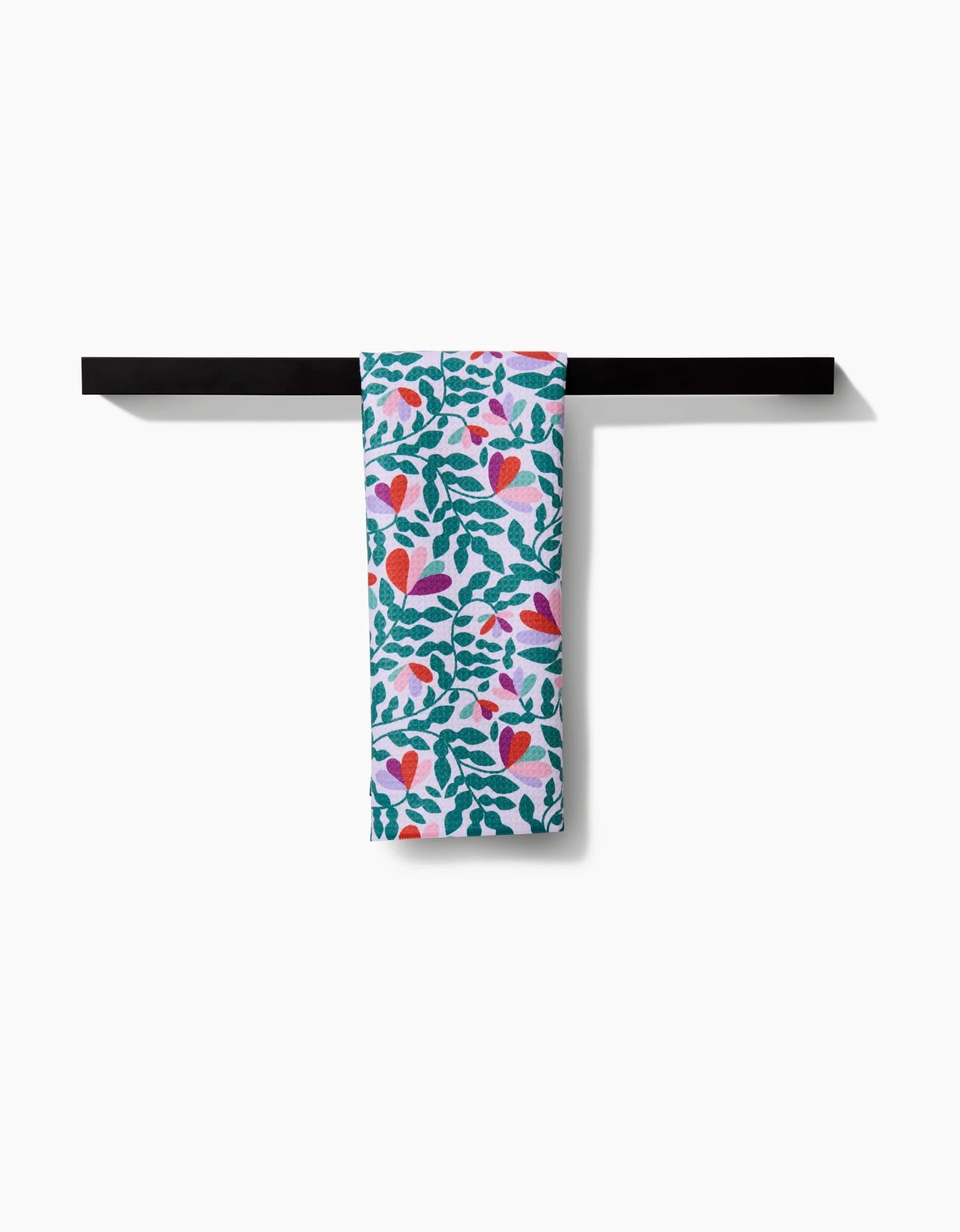 The Spring Wavy Leaves tea towel by Claire Iglesias, crafted from Geoweave fabric, displays green leaves and red-pink heart-shaped flowers, hanging on a black bar against a white background.