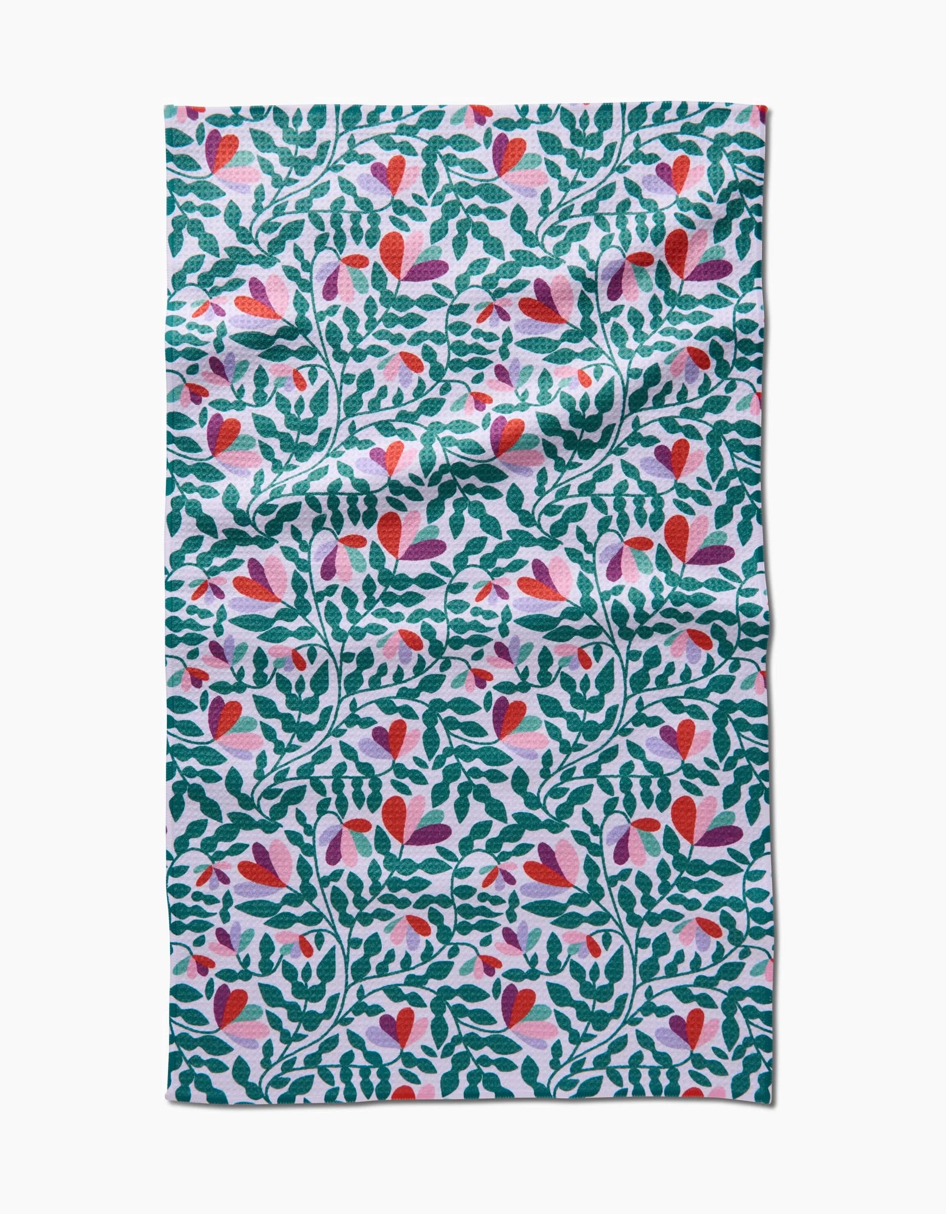 The Spring Wavy Leaves Tea Towel by Claire Iglesias features a vibrant pattern of green leafy branches and pink, red, and purple flowers on white Geoweave fabric. The cloth has a slightly wrinkled texture.