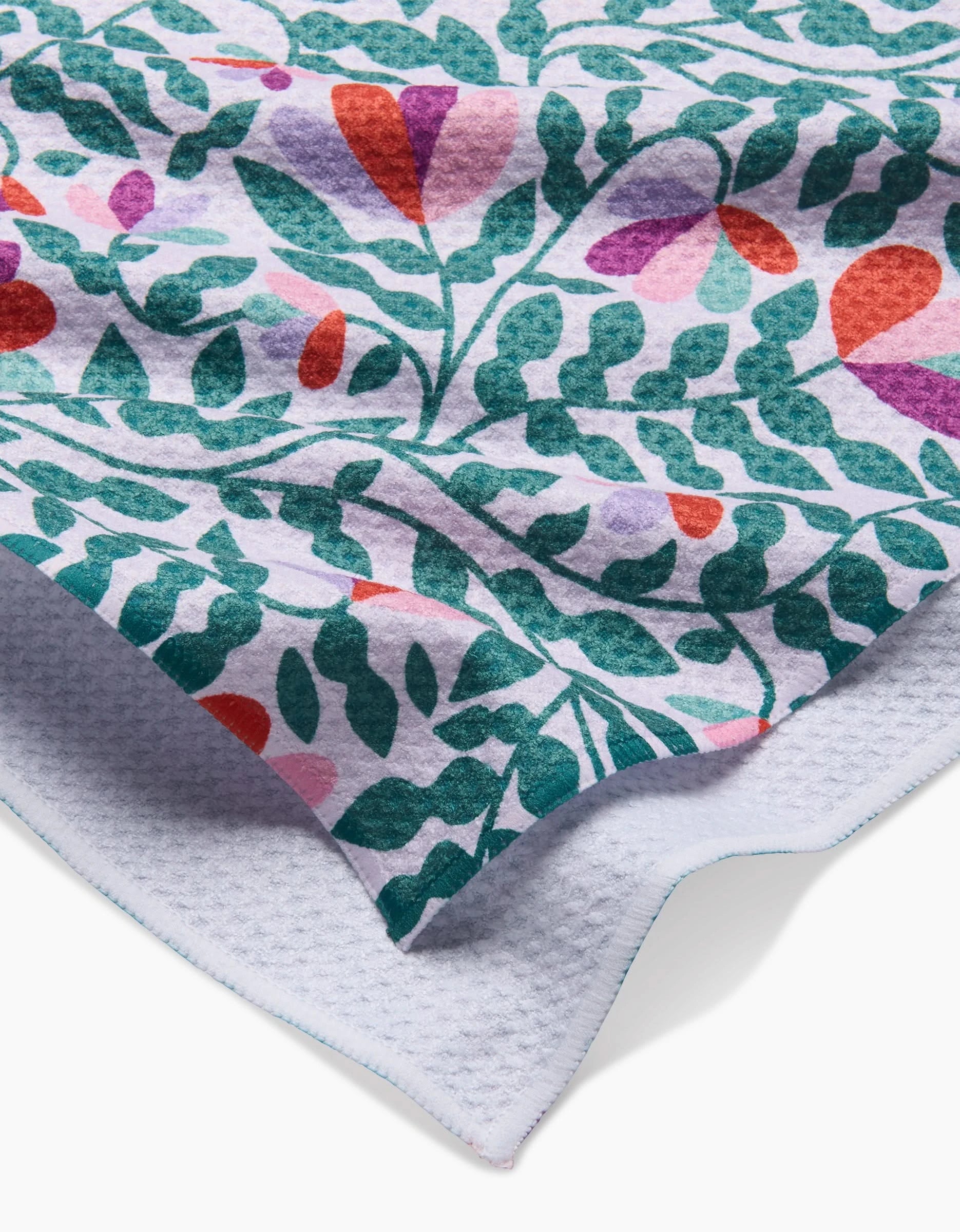 A close-up of the Spring Wavy Leaves Geoweave tea towel by Claire Iglesias, featuring a colorful green, red, purple, and pink floral and leaf pattern on white, with a textured underside and stitched blue edge.