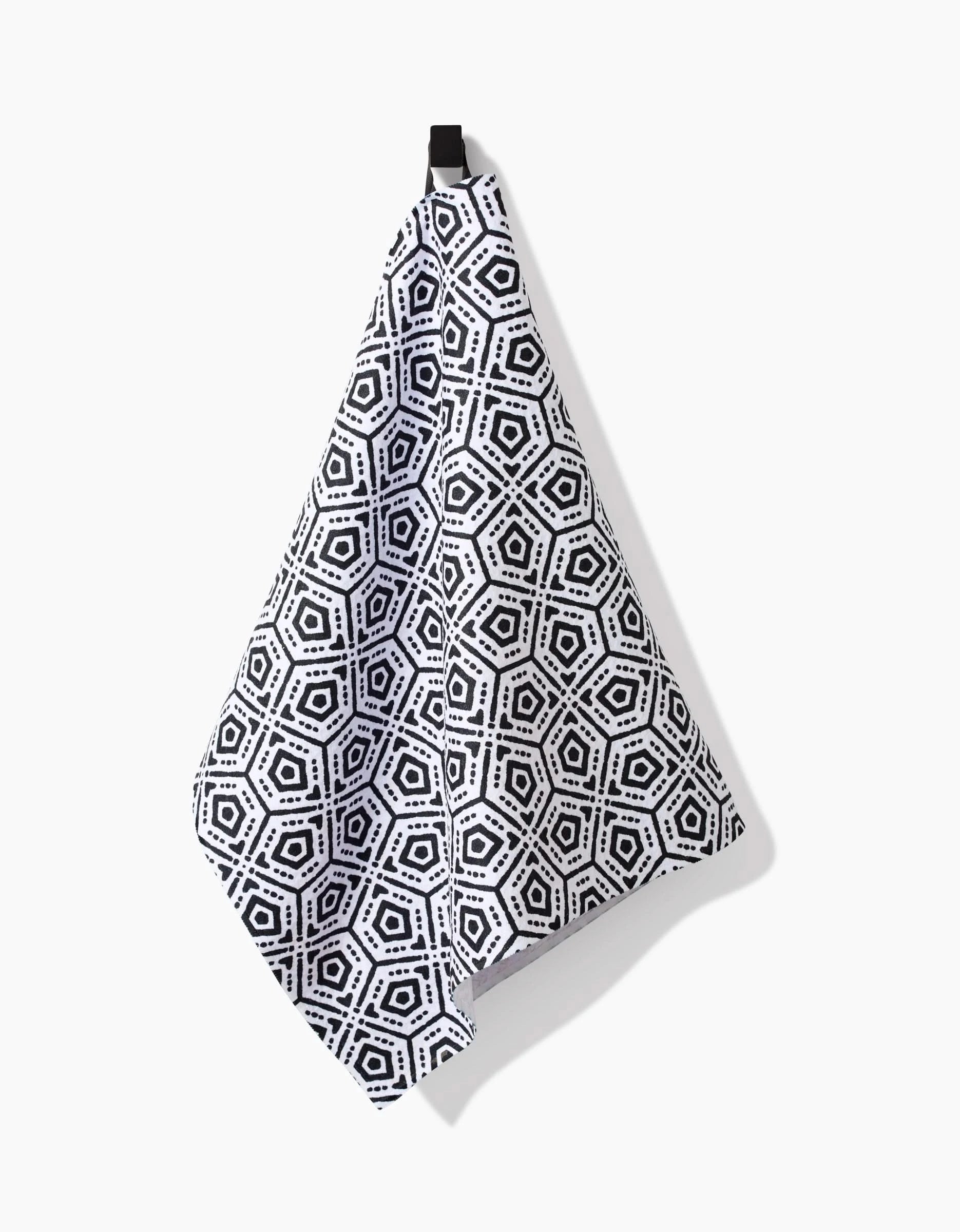 The Squeezebox Tea Towel by Katherine Berkeley, featuring a black and white geometric hexagon pattern in Geoweave™ fabric, hangs from a black loop against a white background.