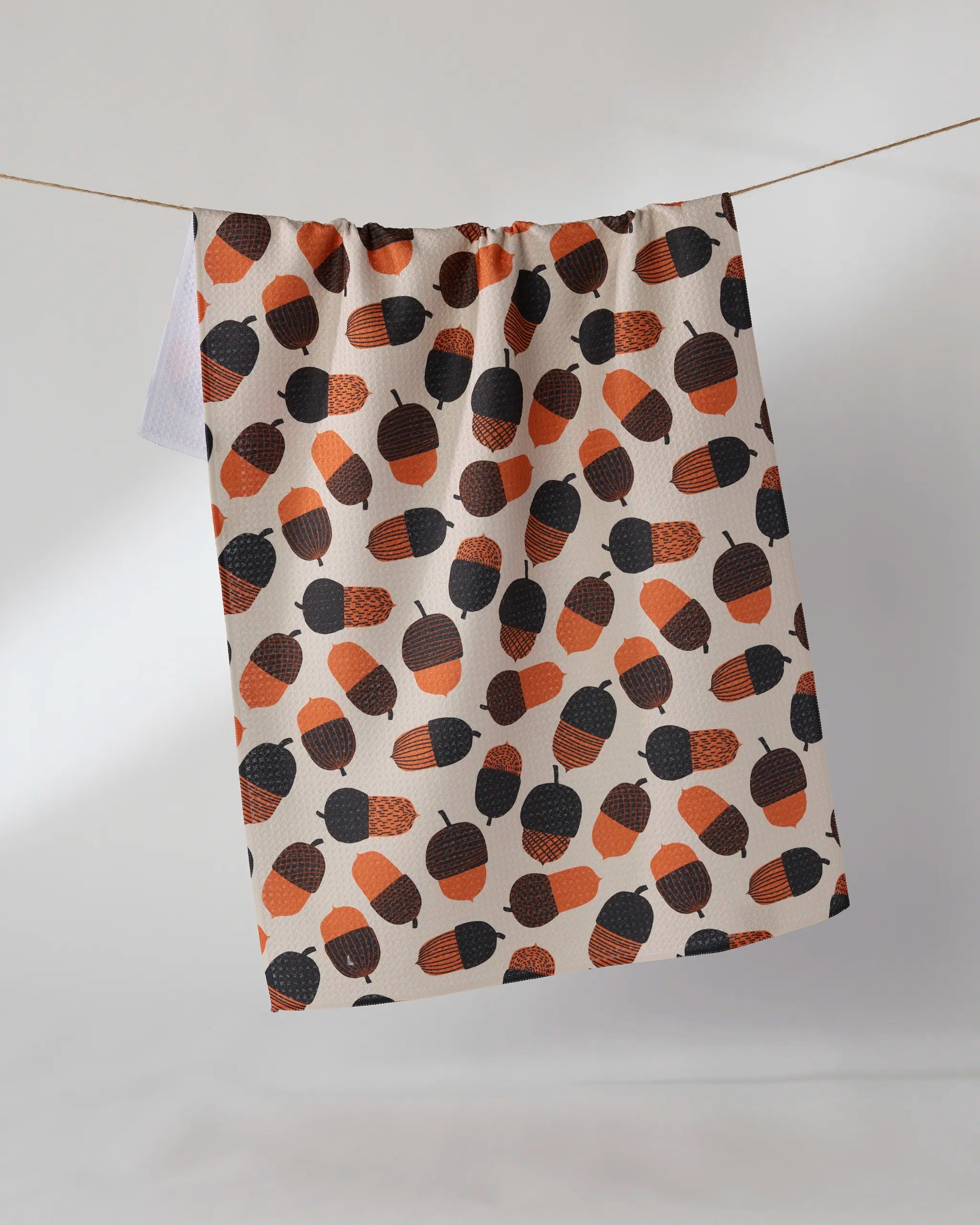 The Geometry "Squirrel Bounty" kitchen towel, made from beige recycled material and featuring a pattern of orange, brown, and black acorns, hangs on a clothesline against a plain light background.