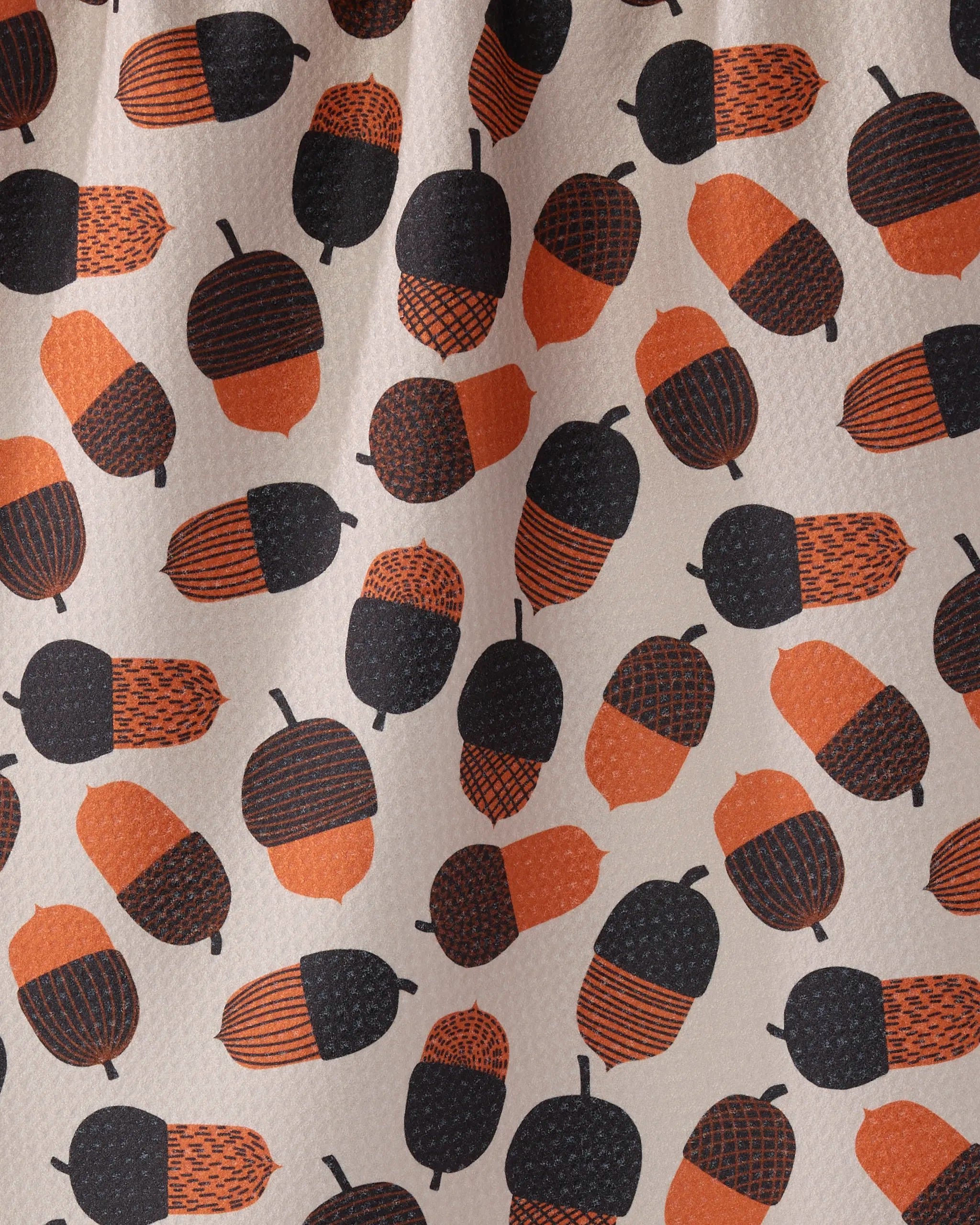 A close-up of Geometry's Squirrel Bounty recycled kitchen towel highlights a repeating pattern of orange and black stylized acorns with varied textures on a light beige background.