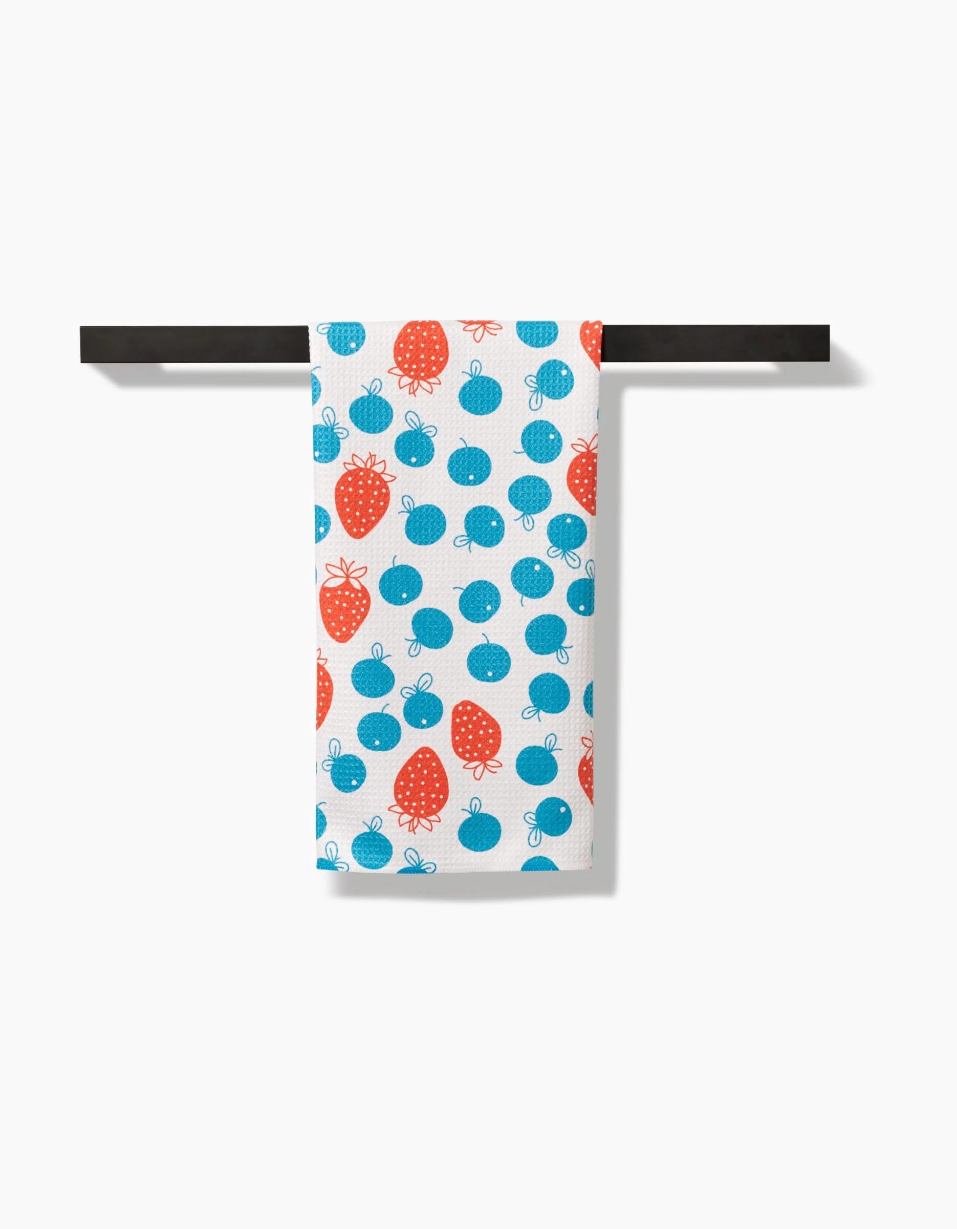 A Sara Mari Star Spangled Berry tea towel, featuring a playful pattern of red strawberries and blue cherries, hangs neatly on a black horizontal towel bar against a plain white background.