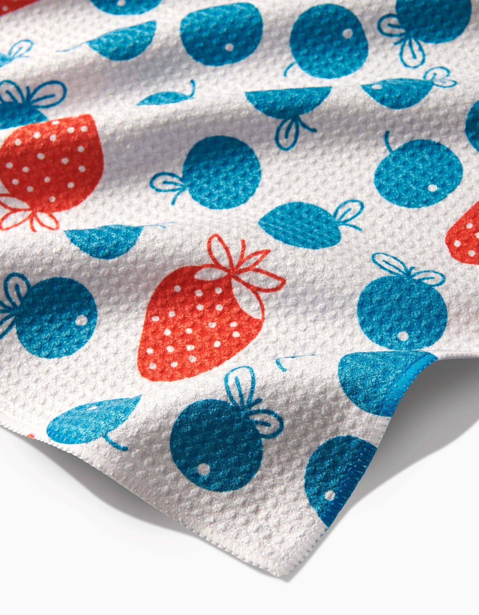 Close-up of the Sara Mari Star Spangled Berry tea towel, made from Geoweave fabric, featuring a textured white cloth with red strawberries and blue cherries adorned with small white polka dots scattered across the surface.