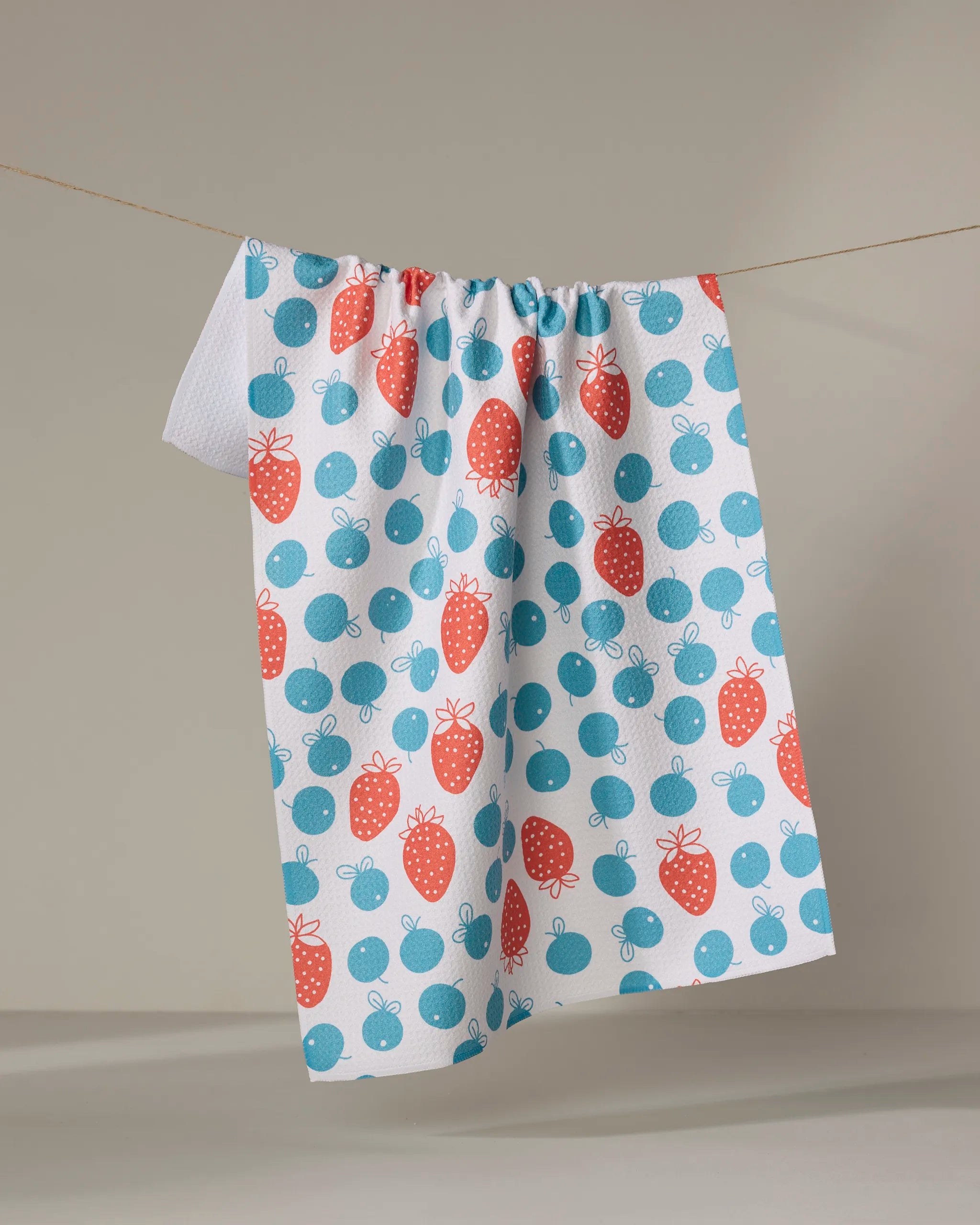 The Sara Mari "Star Spangled Berry" tea towel, featuring a pattern of red strawberries and blue cherries, hangs on a clothesline against a neutral background.