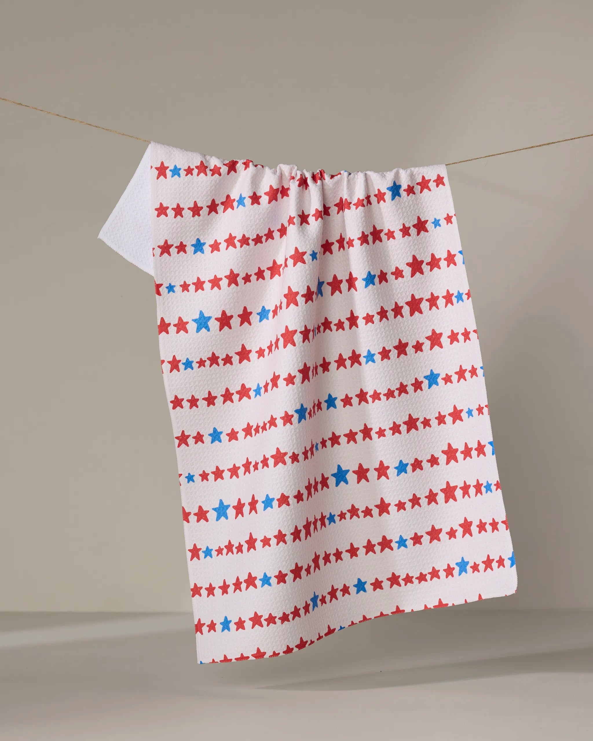 The Kayla Ann "Starry Stripes" eco-friendly white kitchen towel, featuring red and blue stars and stripes, hangs on a clothesline against a simple light background.