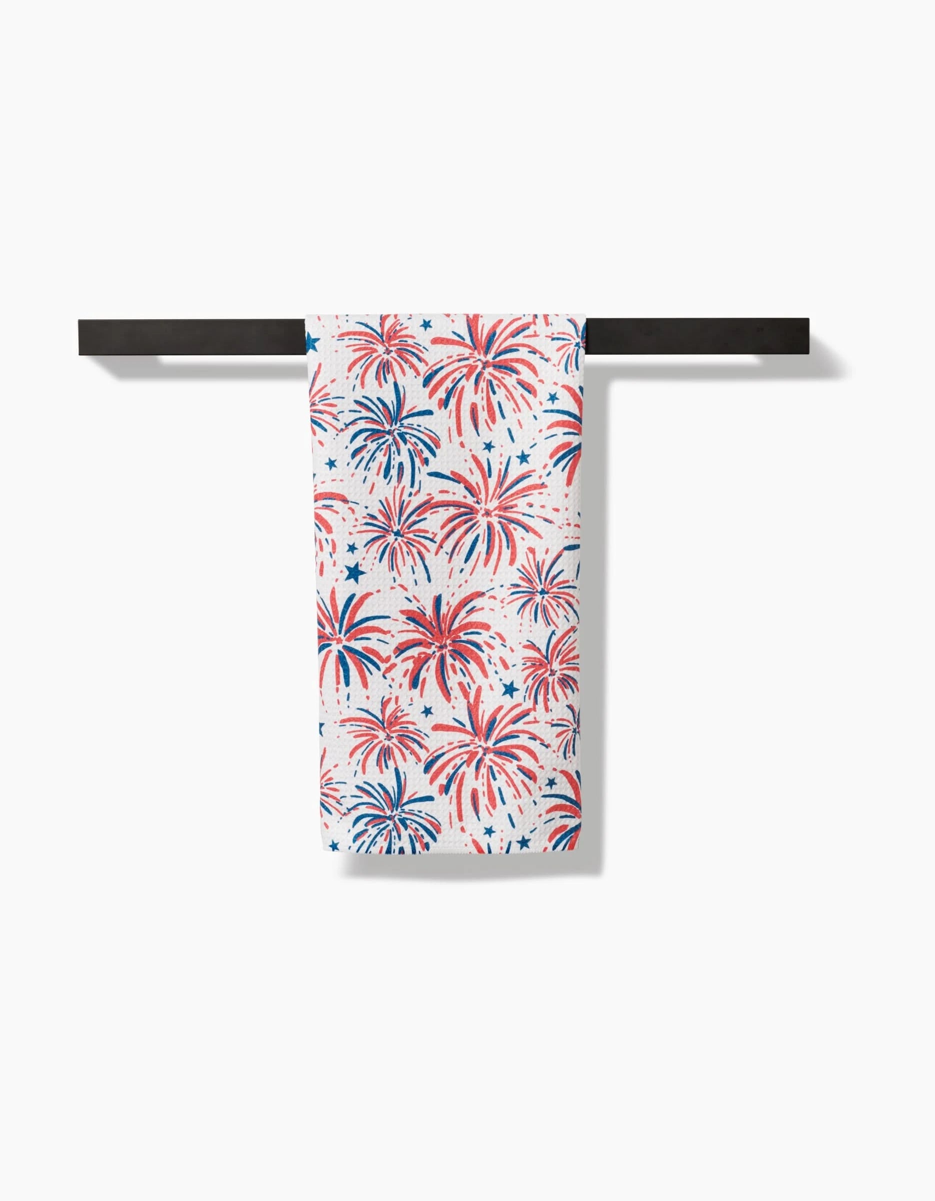 The Stars And Fireworks tea towel by Hufton Studio, featuring a red and blue fireworks pattern on Geoweave fabric, hangs neatly on a black towel bar against a white background.