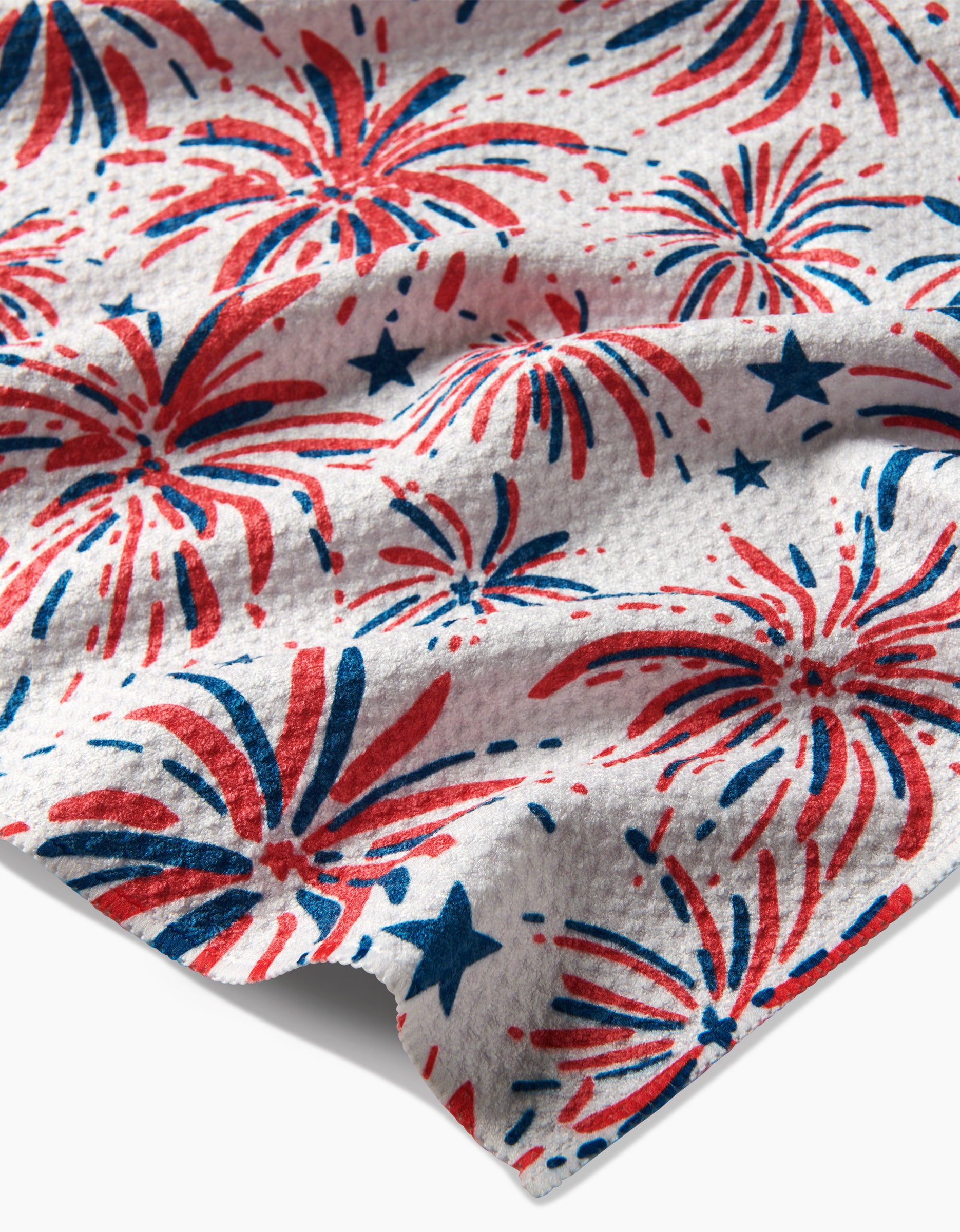 A close-up of the Hufton Studio Stars And Fireworks Geoweave Tea Towel shows red and blue fireworks and stars on white recycled fabric, with a textured, slightly wrinkled surface creating subtle shadowed folds.