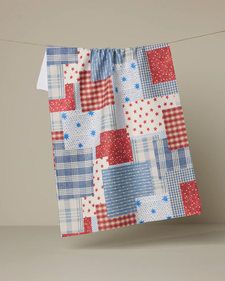 The Geometry Stars and Stitches recycled kitchen towel, featuring a red, white, and blue patchwork design with stars, checks, and plaid patterns, hangs on a clothesline against a neutral background.