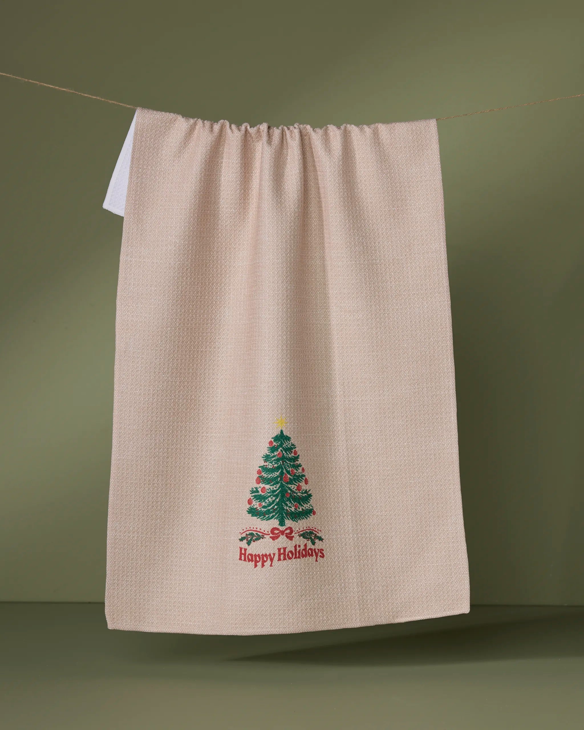 The Geometry Starwood tea towel, made from recycled materials, hangs on a line against a green background, showcasing an embroidered Christmas tree with “Happy Holidays” in red below.