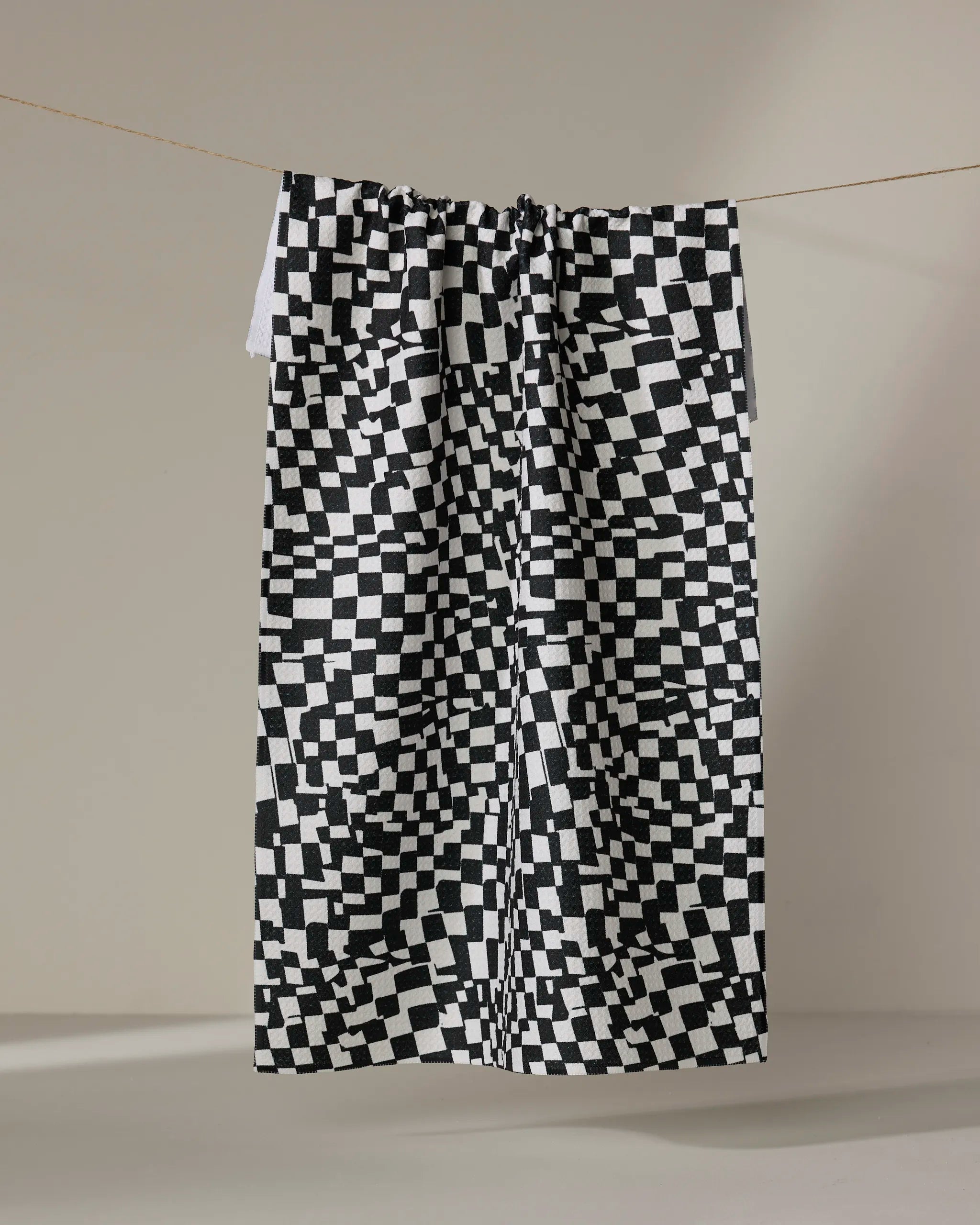 The Geometry Static kitchen towel, featuring a black and white pixelated checkered pattern, hangs from a string against a plain light background.