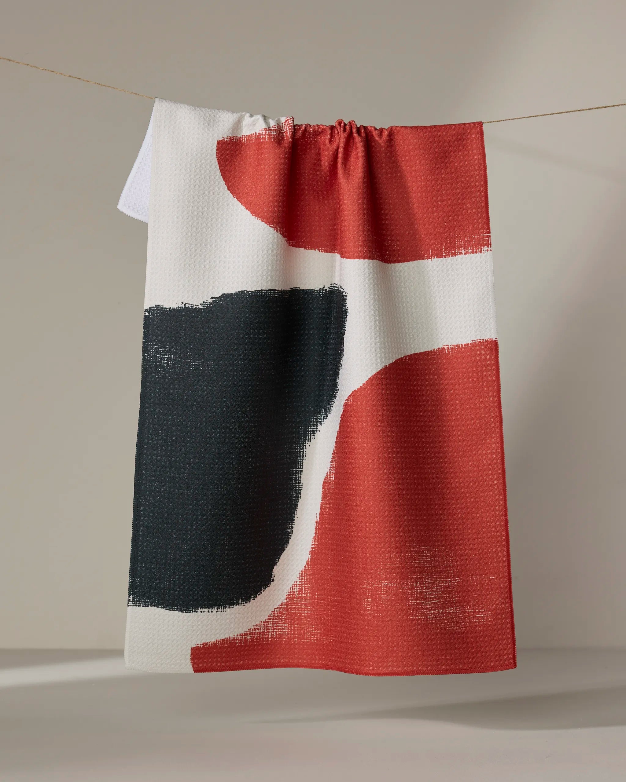 The Geometry "Still" tea towel, featuring an abstract design in red, black, and white with bold curved shapes and brushstroke textures, dries quickly and hangs on a clothesline against a neutral background.
