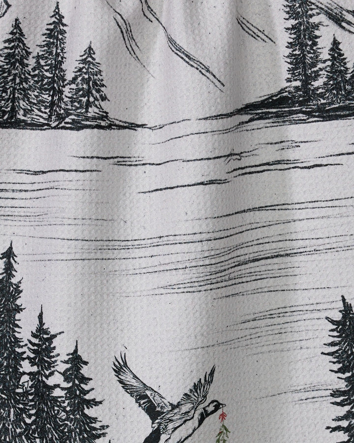 The Stillpine tea towel by Geometry features a black and white mountain landscape with pine trees, a lake, and a bird carrying a branch—crafted from recycled materials and perfect for your kitchen.
