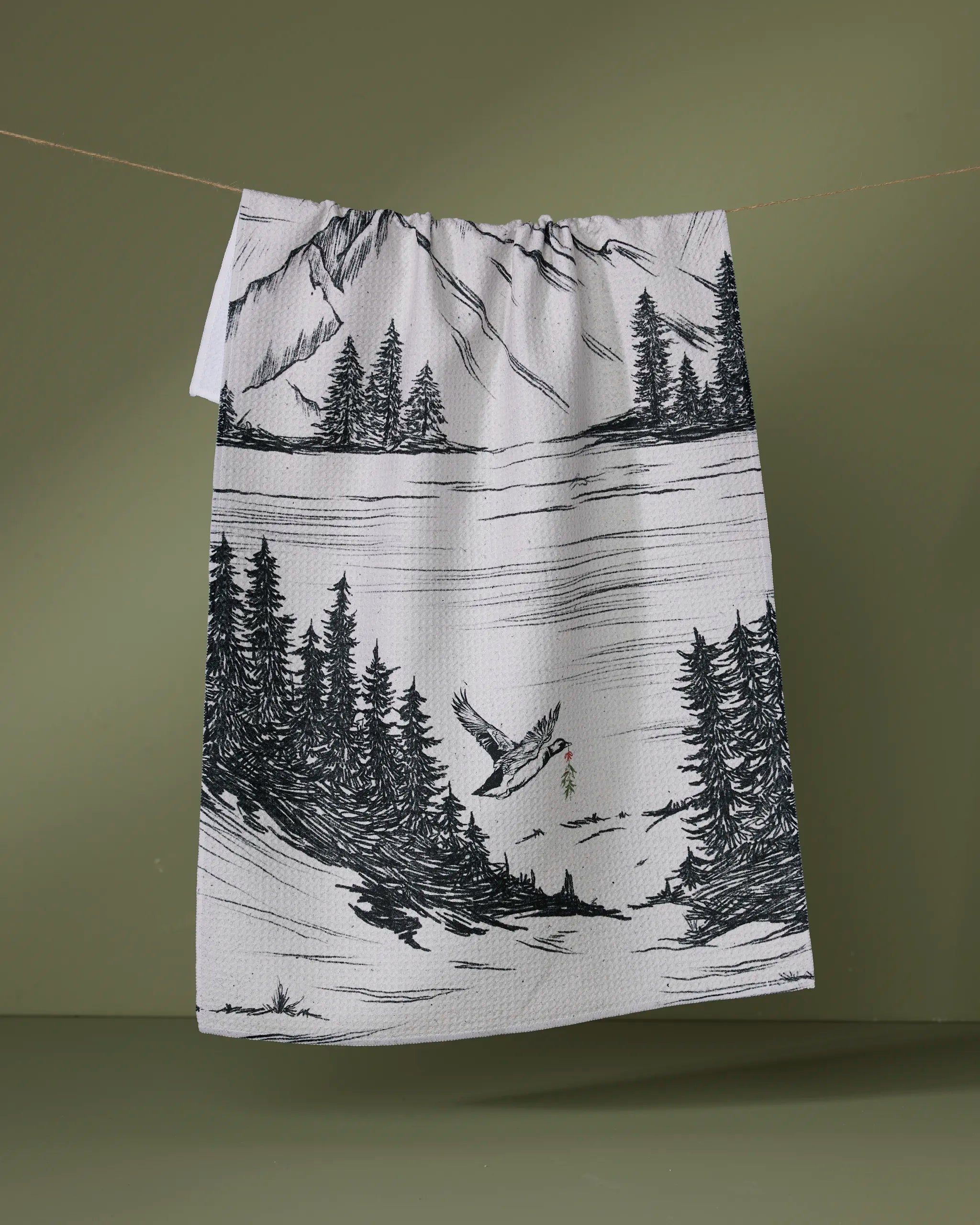 The Geometry Stillpine tea towel features ultra-absorbent fabric and a black-and-white print of forests, mountains, and a soaring bird, set against an olive green background.