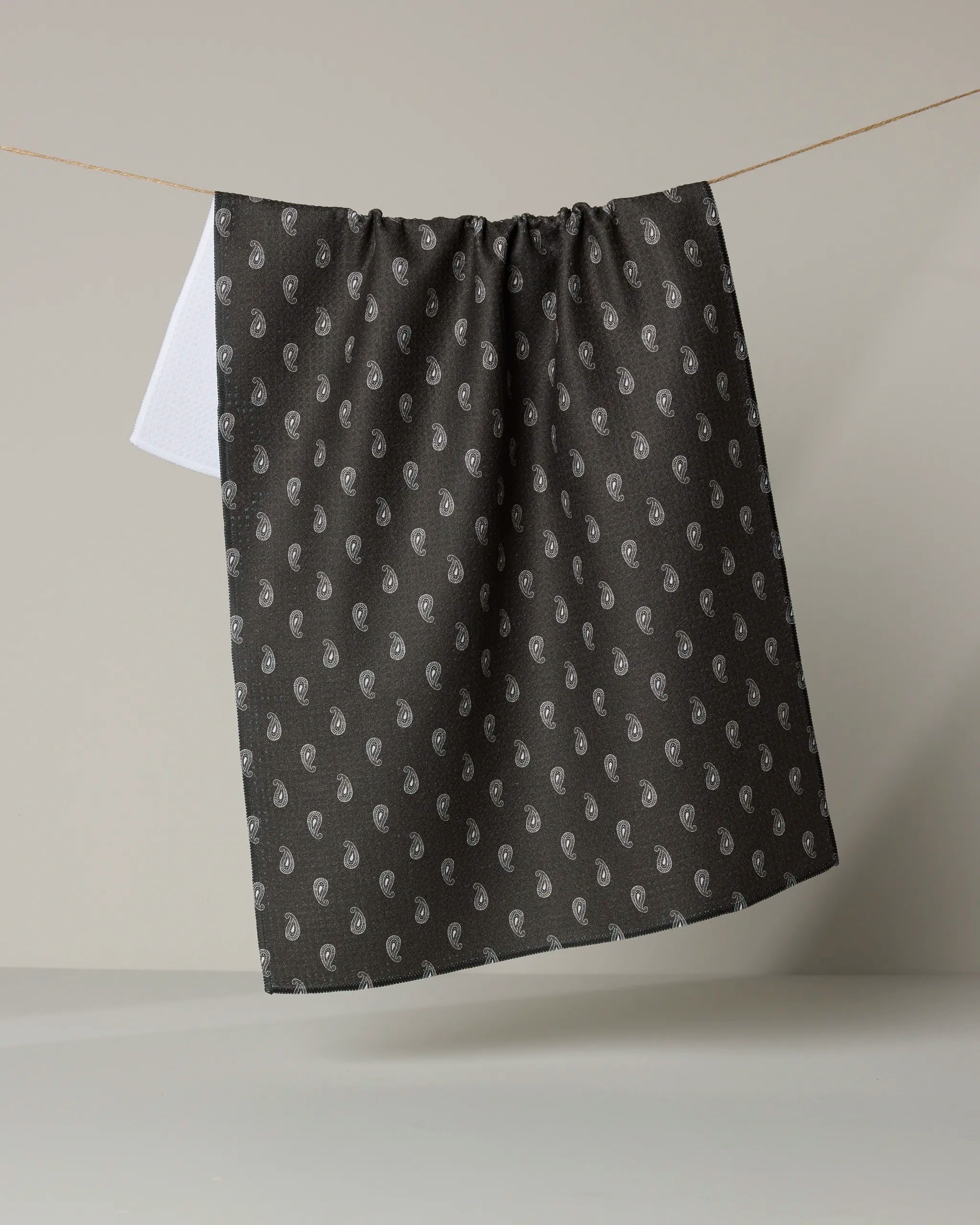 The Geometry Stirrup Espresso recycled kitchen towel, featuring a small white paisley pattern on black, hangs from a string against a light gray background with part of a white tea towel visible behind it.