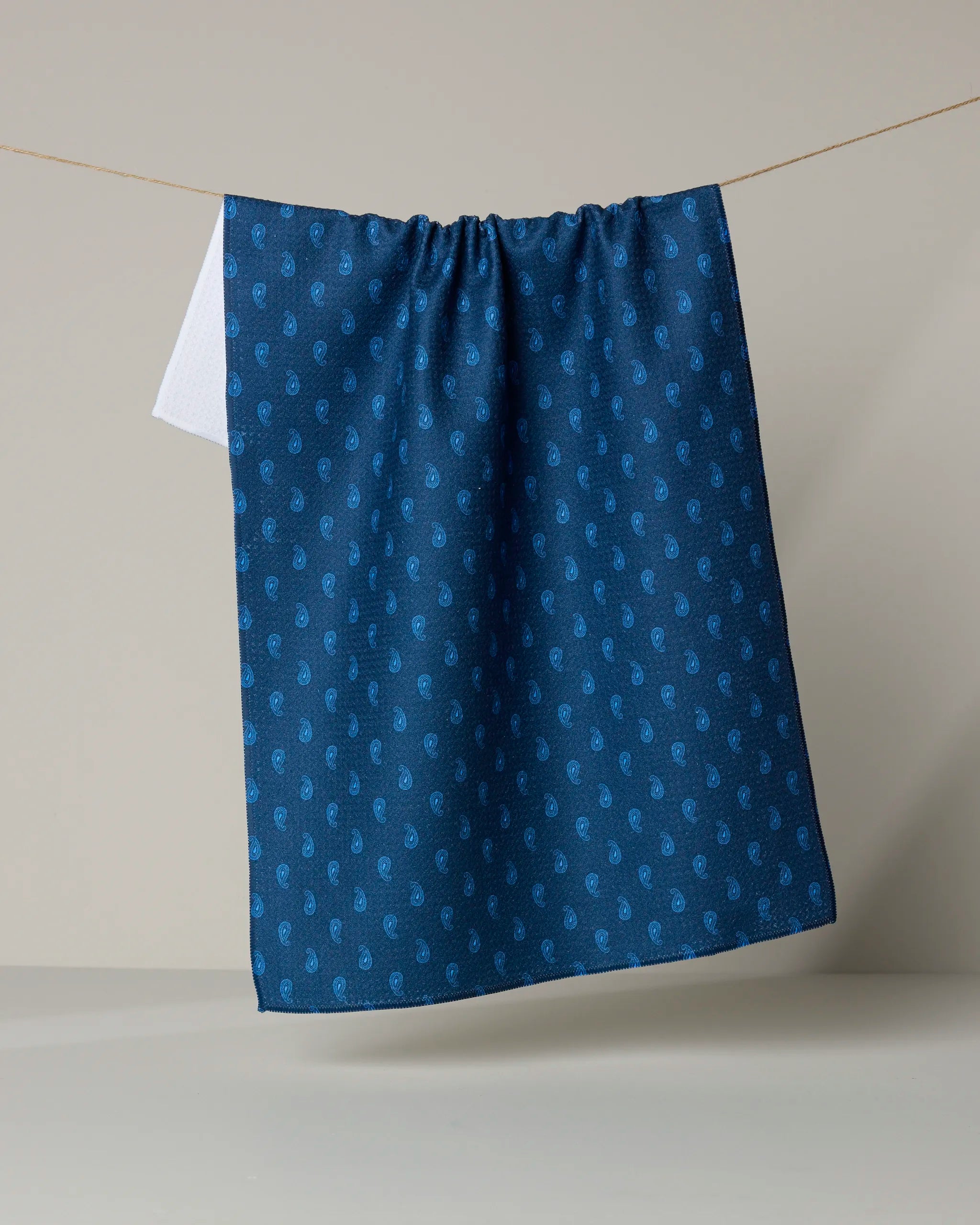 The Stirrup Indigo kitchen towel by Geometry, featuring a dark blue hue with a subtle repeating light blue paisley pattern, hangs on a clothesline against a neutral background.
