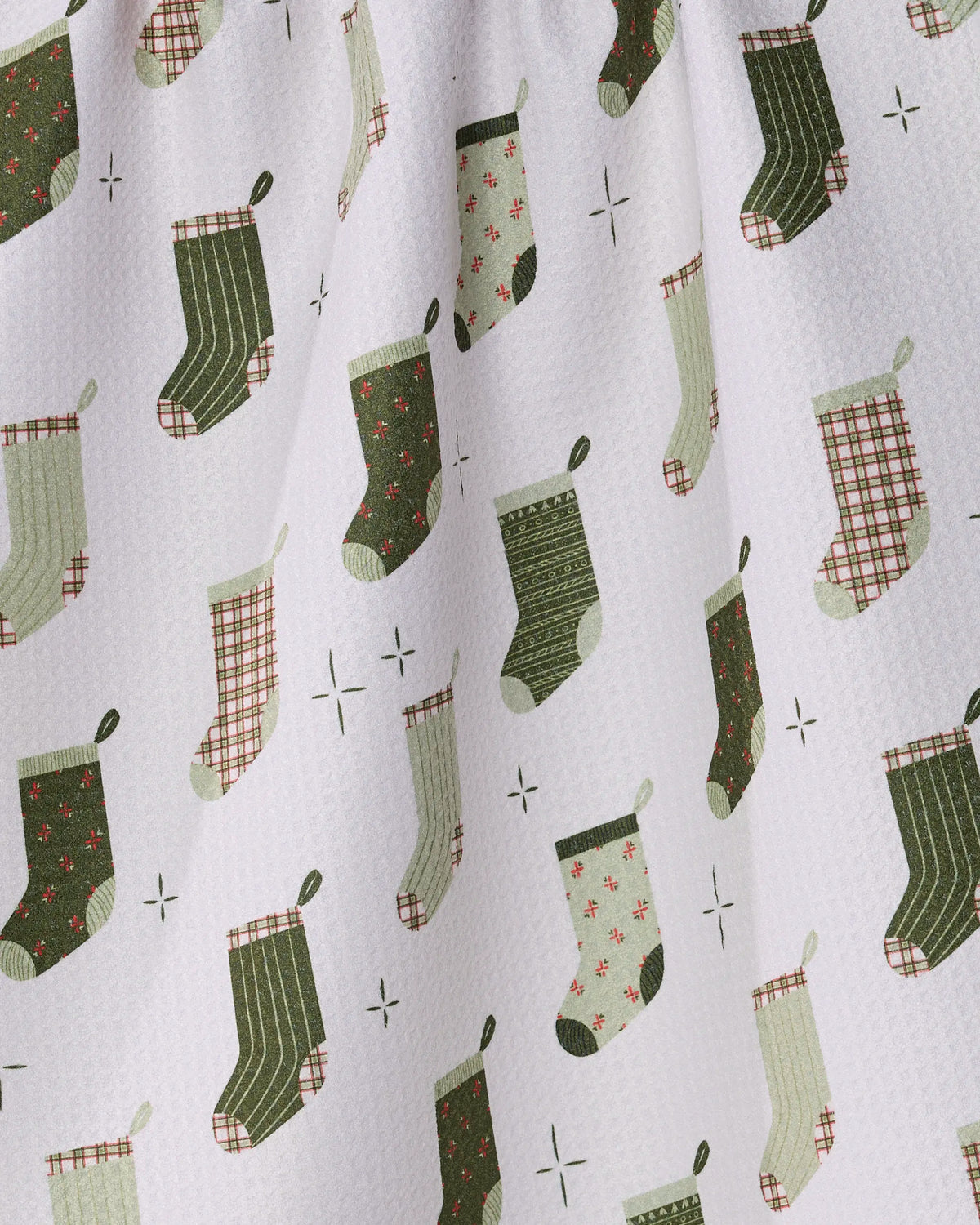 The Stocking Medley tea towel by Geometry features a white fabric adorned with a repeating pattern of green and red Christmas stockings—each showcasing plaid, striped, or polka dot designs—alongside small star shapes. Gathered at the top and crafted from recycled materials, this charming piece adds an eco-friendly touch to your holiday kitchen decor.