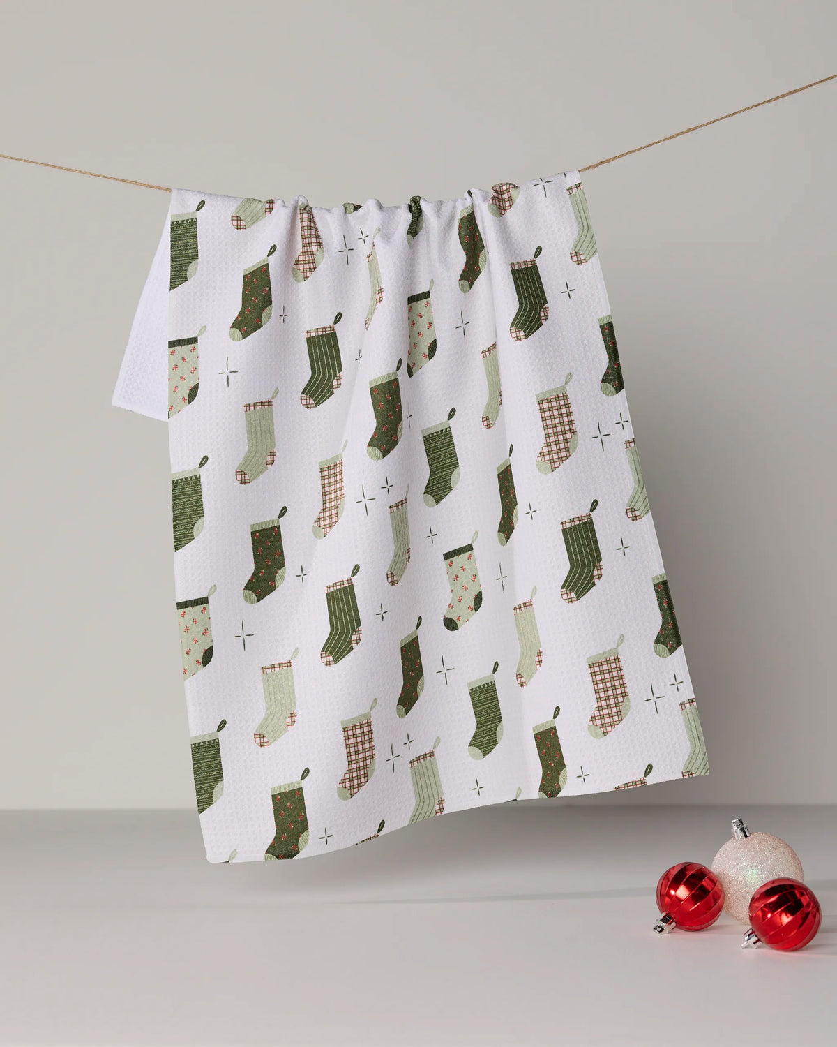 A Geometry Stocking Medley towel, featuring ultra-absorbent white fabric adorned with green and beige Christmas stocking patterns, hangs on a string. Two red and one silver Christmas ornaments are displayed on the gray surface below.