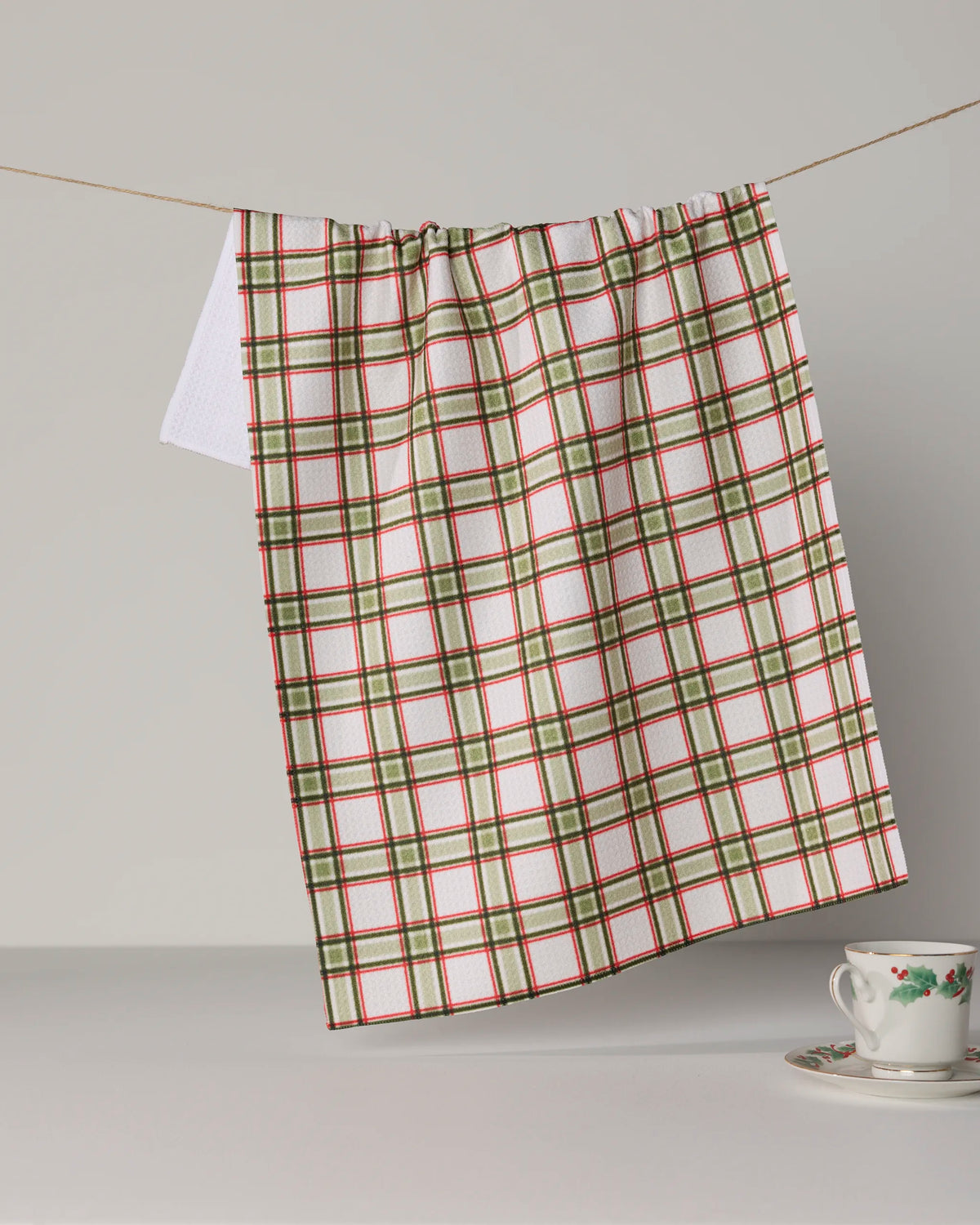A Stocking Plaid tea towel by Geometry, featuring green, red, and white plaid and crafted from recycled materials, hangs on a string. Below it, a white teacup with a floral pattern rests on a saucer against a neutral background.